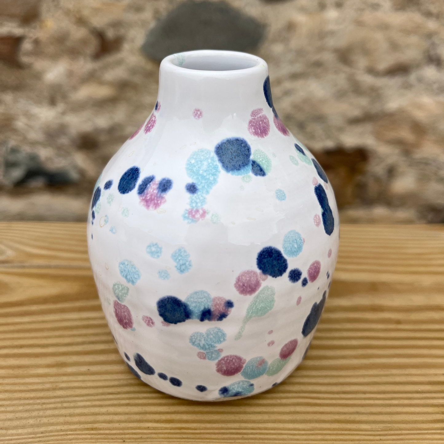 White vase with colorful dot patterns on a wooden surface with a stone wall background