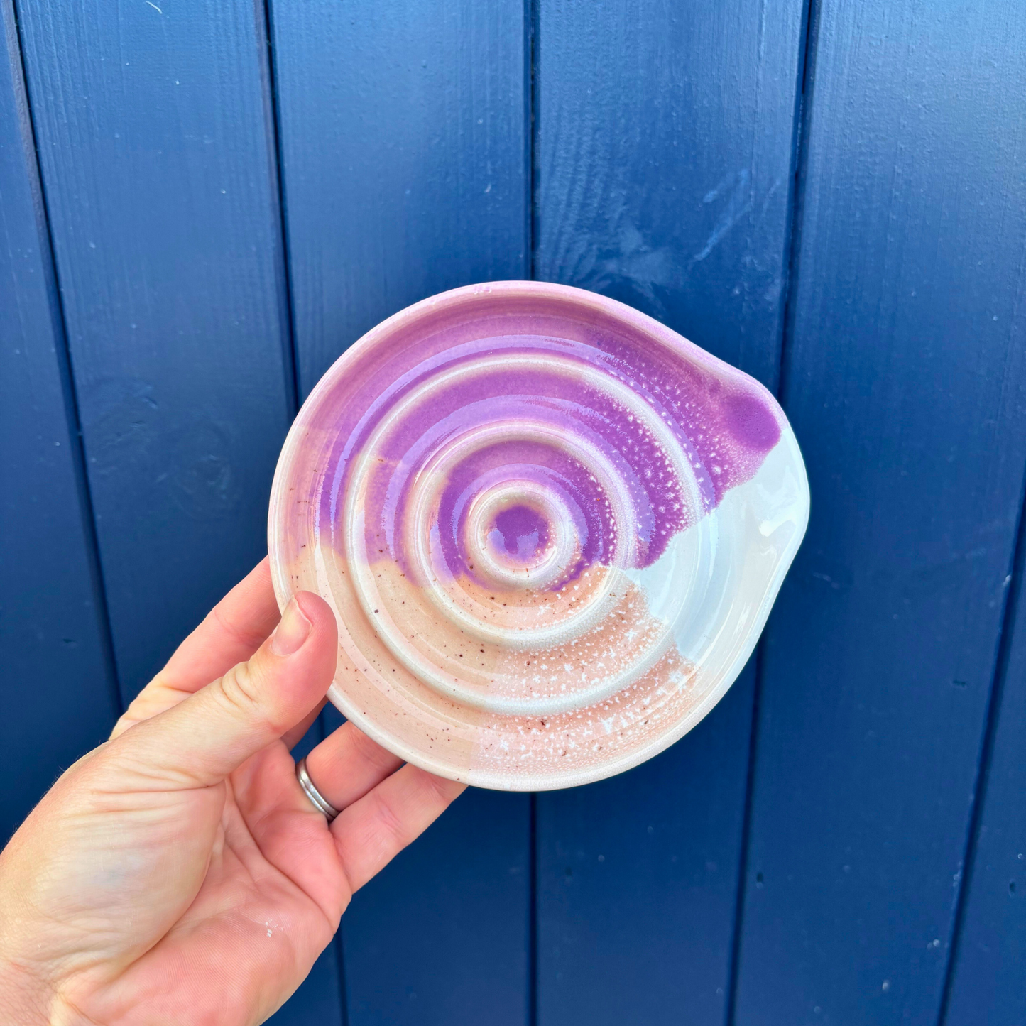 Hand holding a ceramic spoon rest with purple and white spiral design against a blue wooden background