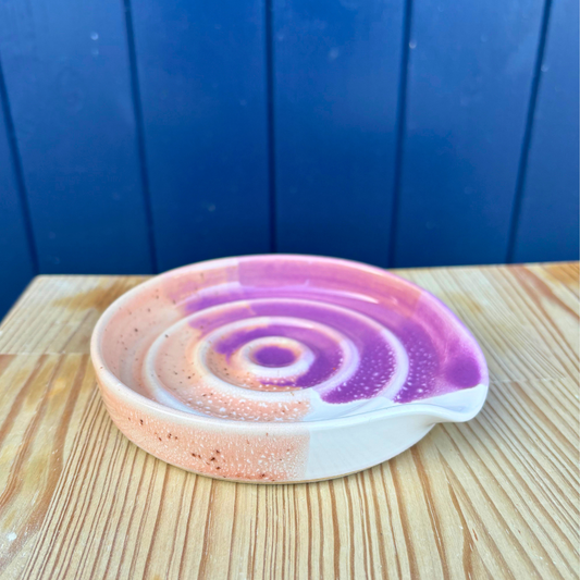 Ceramic spoon rest dish with purple and pink spiral design on a wooden surface with a blue background
