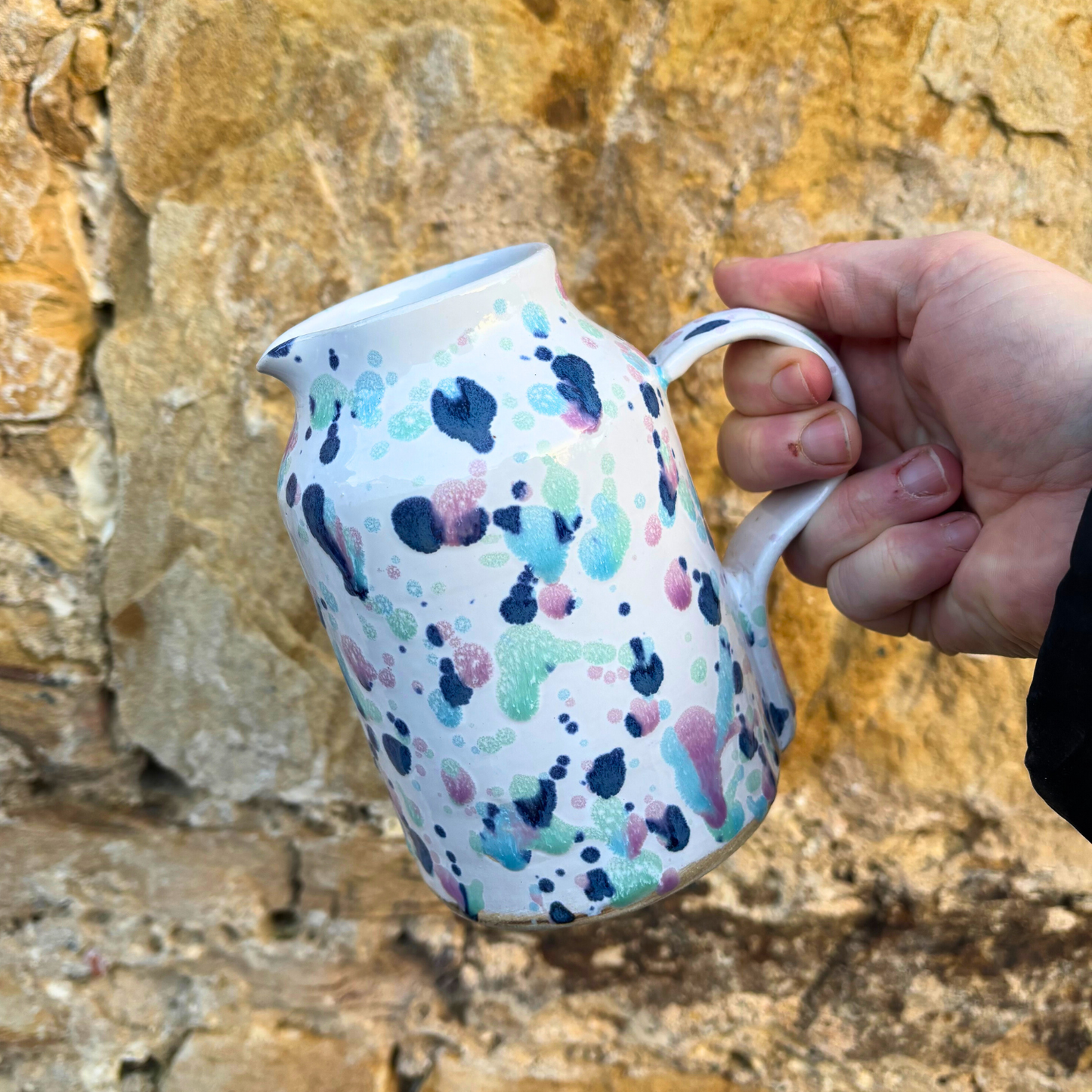 Hand holding a ceramic pitcher with colorful abstract design against a stone wall background