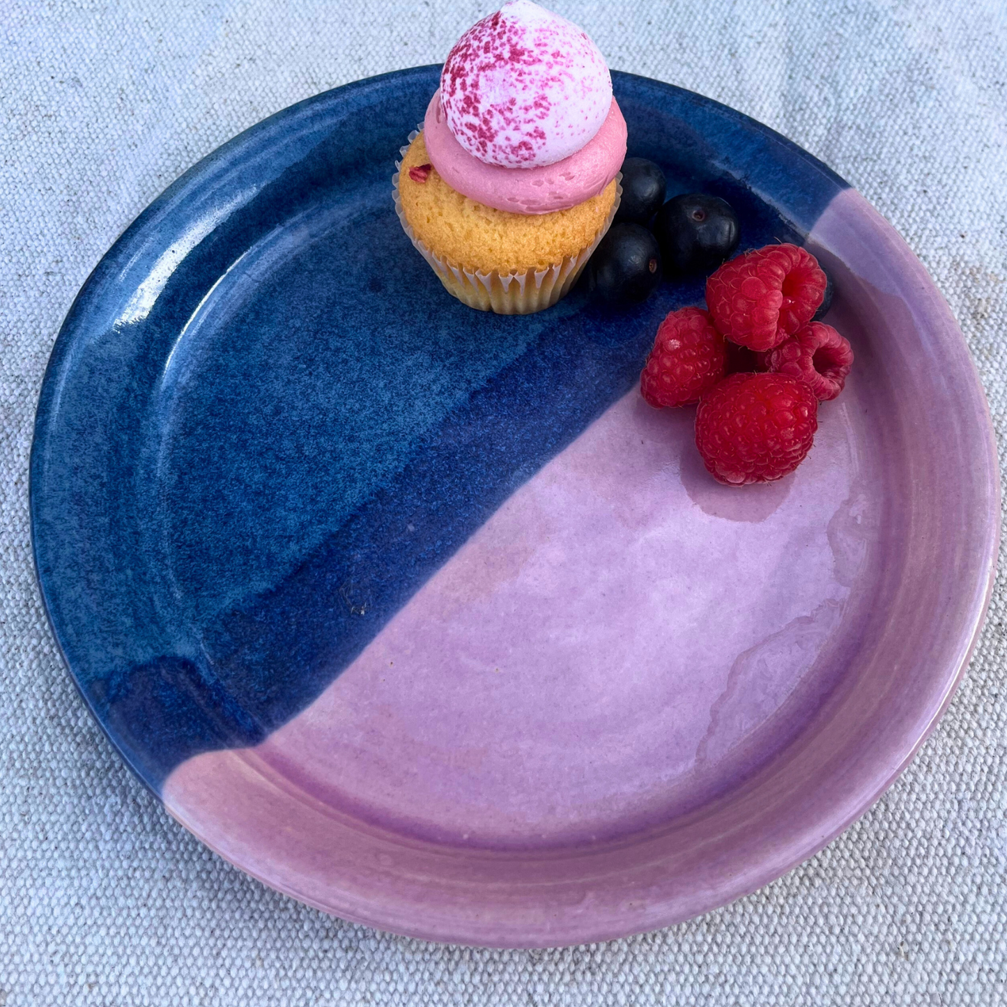 Cupcake with pink frosting and berries on a half blue, half purple ceramic plate.