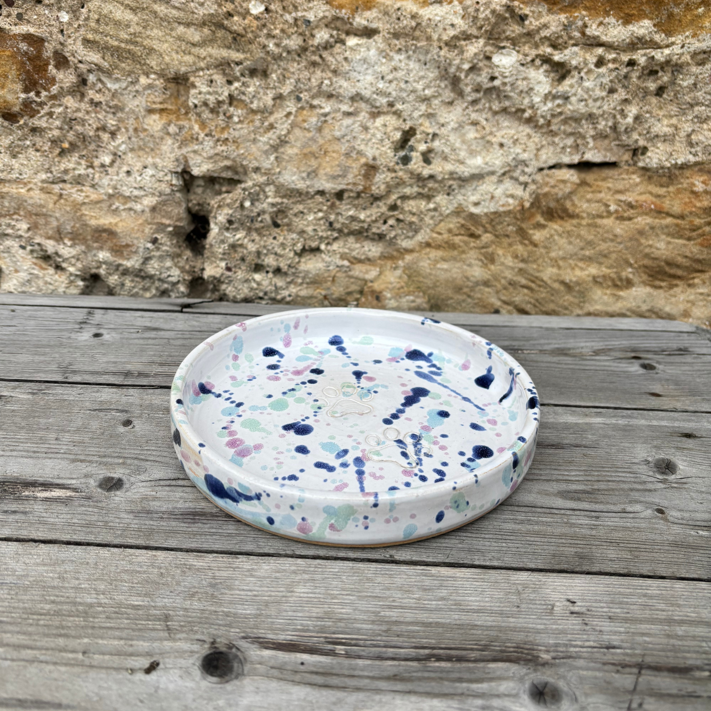 Round ceramic plate with multicolored speckled design on a wooden surface with stone wall background