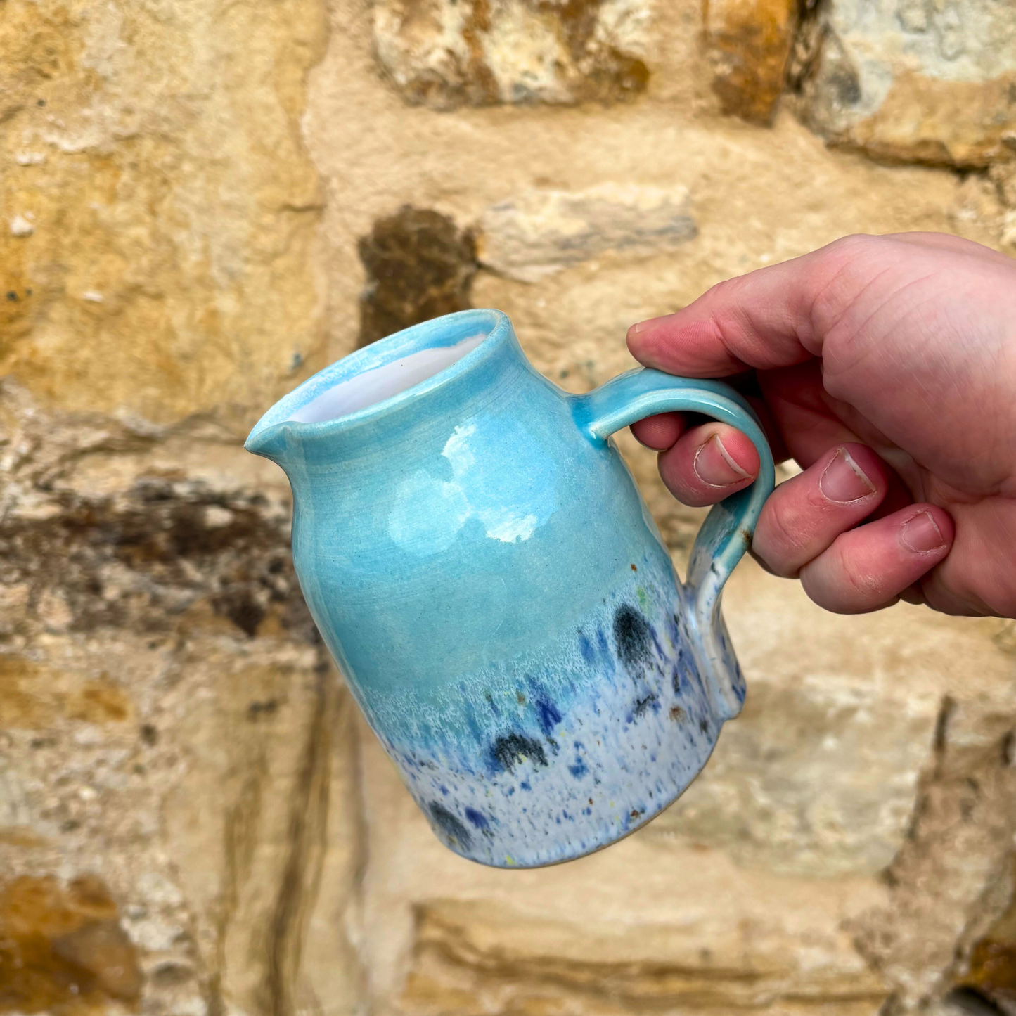 Hand holding a blue ceramic pitcher against a stone wall background