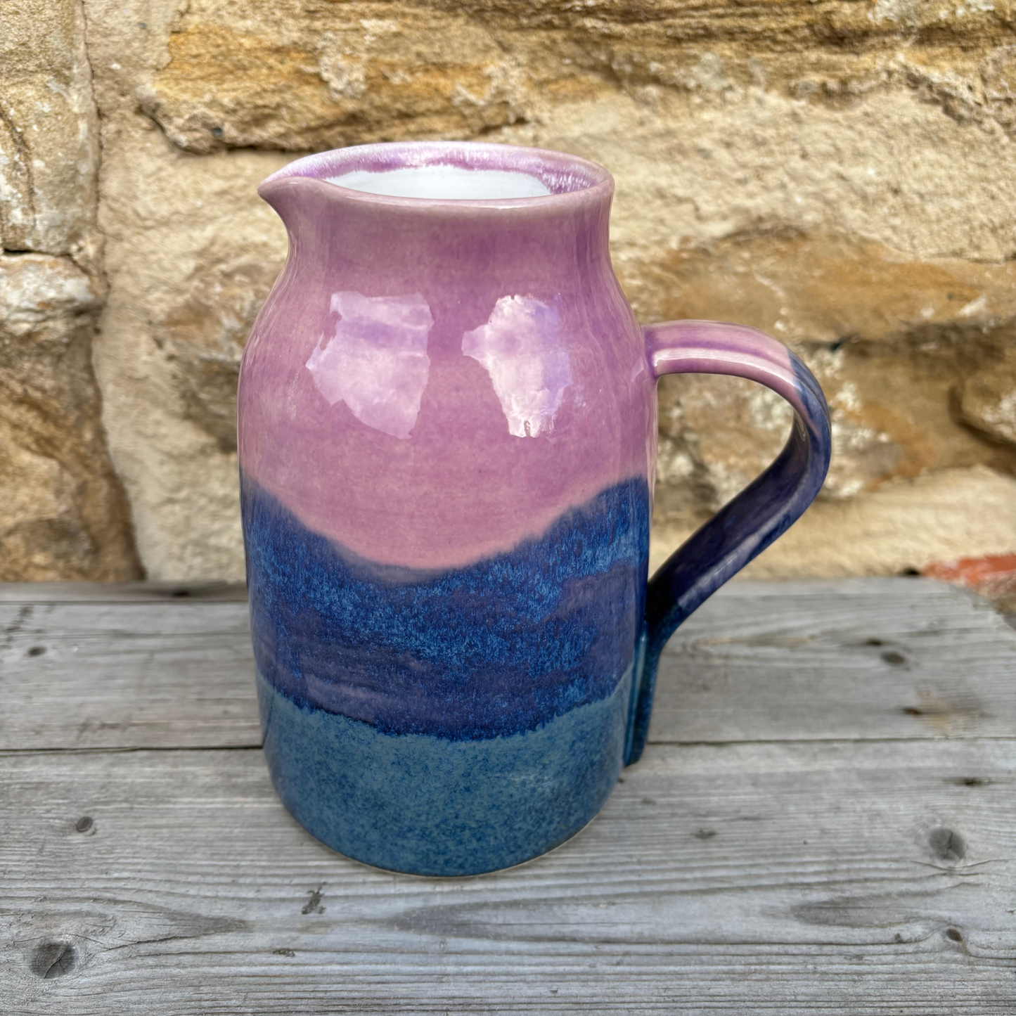 Ceramic pitcher with purple and blue gradient on a wooden surface with stone wall background