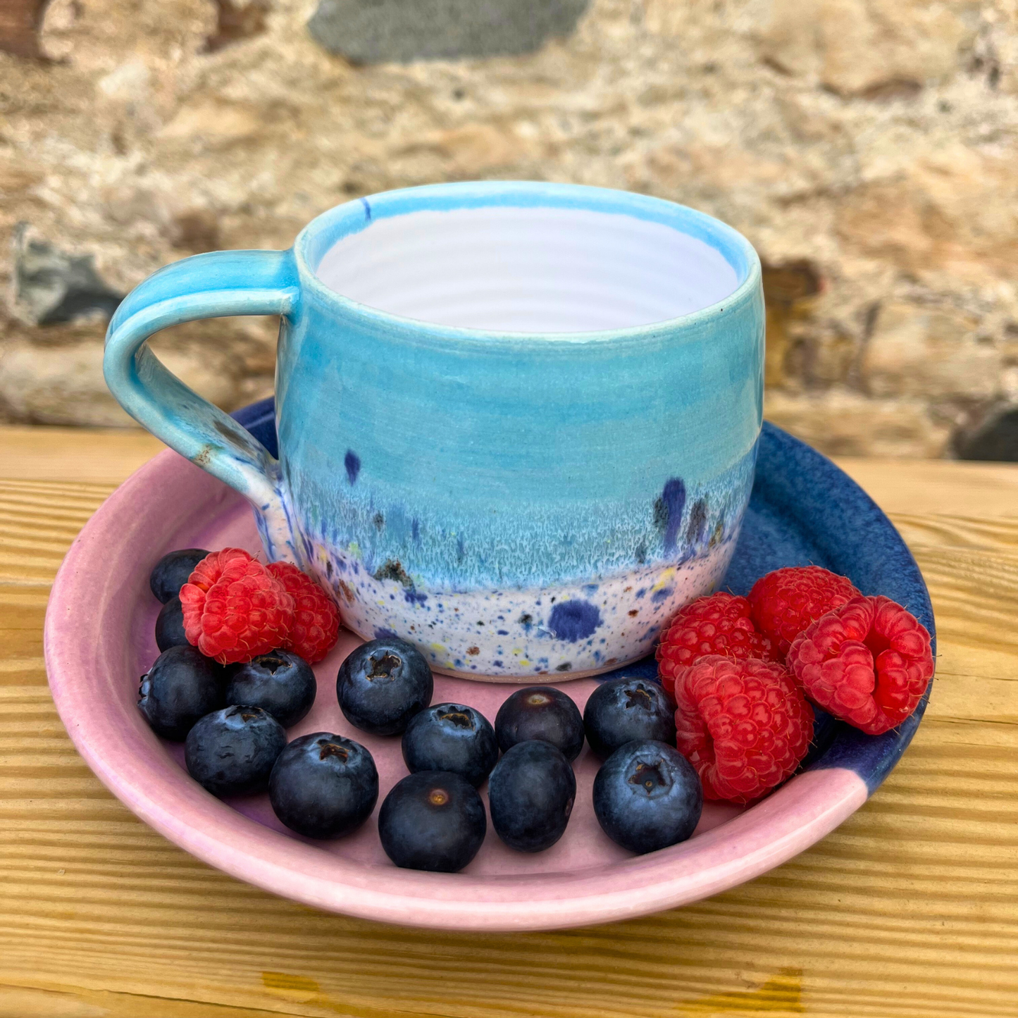 Blue ceramic mug with speckled design on a pink plate with blueberries and raspberries, against a stone wall background.