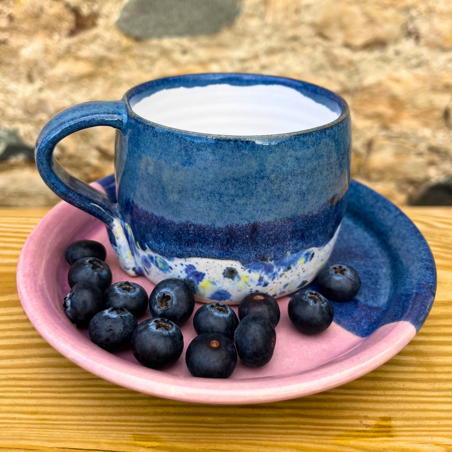 Blue ceramic cup and saucer with pink rim on a wooden surface, containing blueberries.