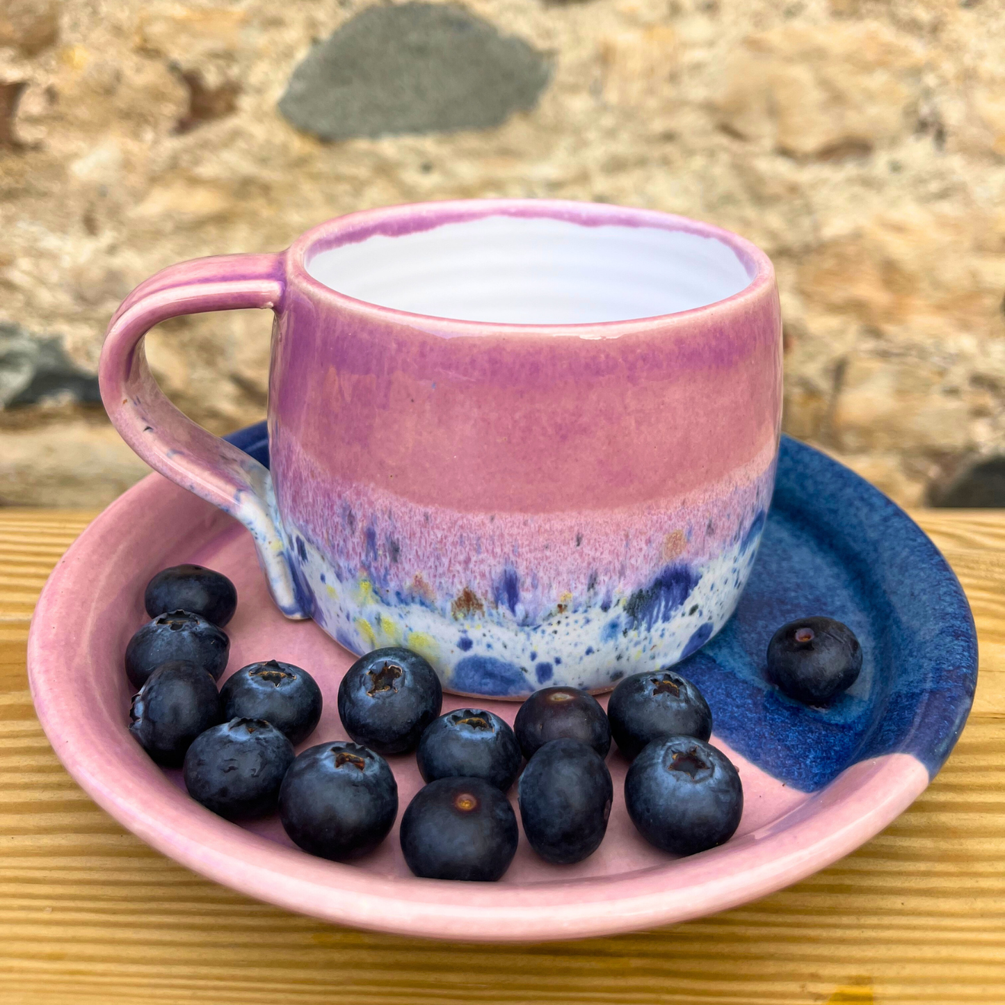 Pink ceramic mug with blueberries on a matching pink saucer against a natural stone background