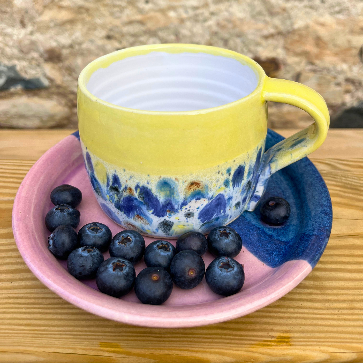 Yellow ceramic mug with blue and white abstract design on a pink ceramic plate with blueberries, set against a natural stone background.