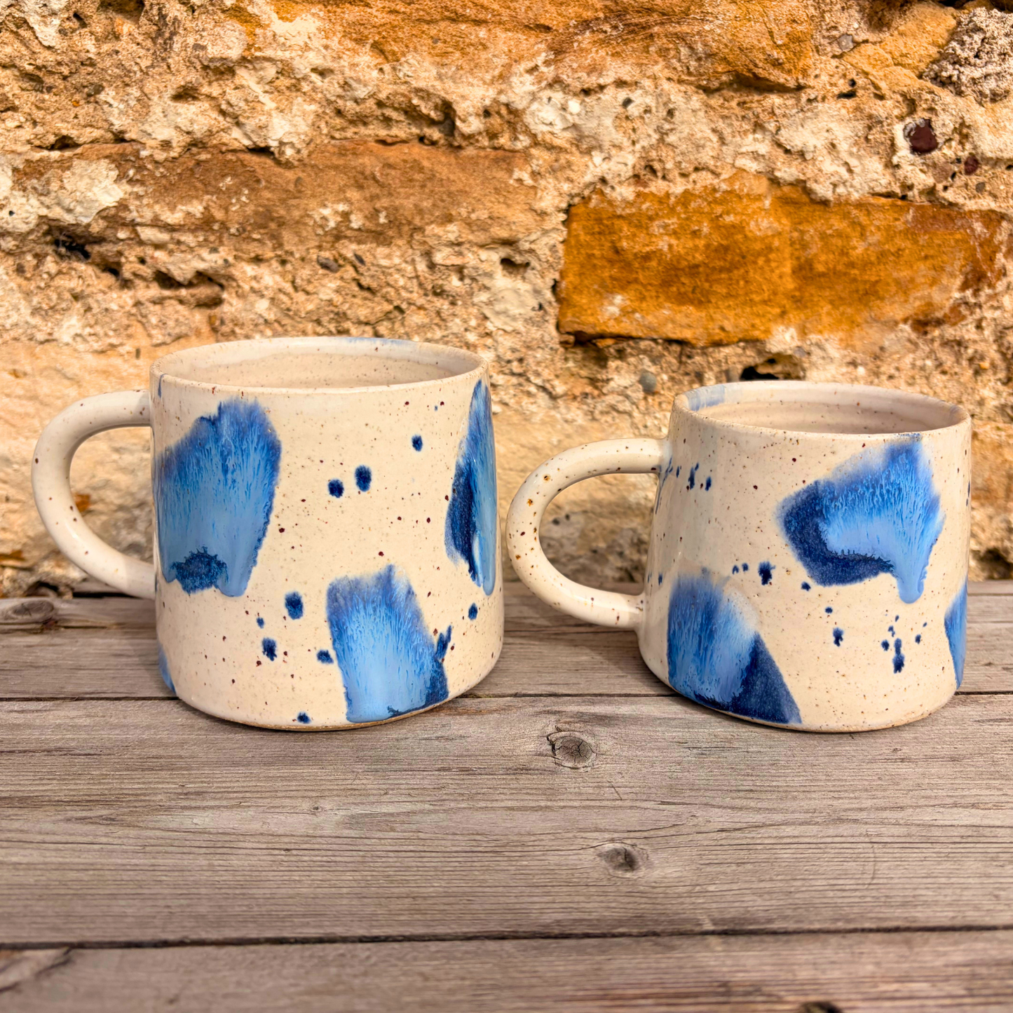 Two ceramic mugs with blue abstract patterns on a wooden surface with a stone wall background.