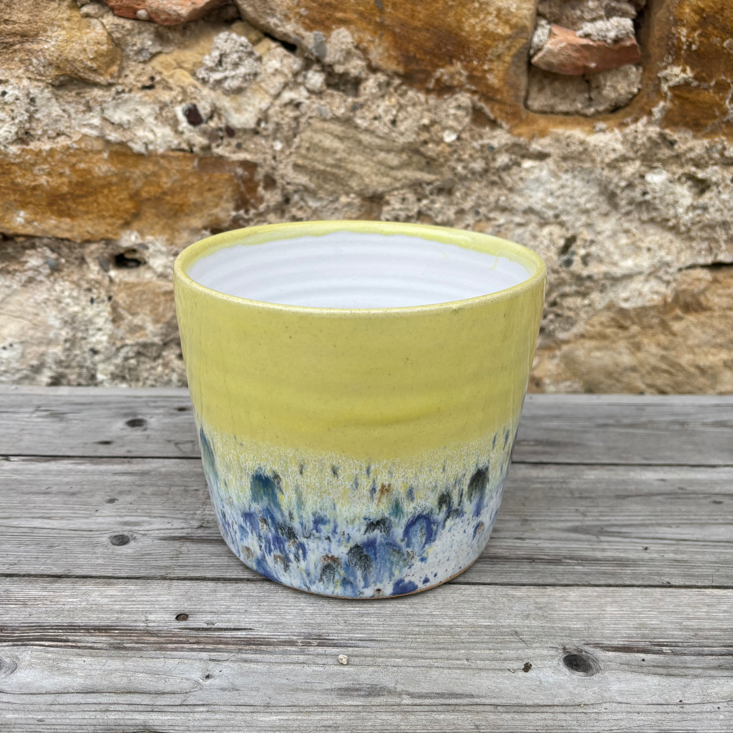 Yellow ceramic pot with blue speckled design on a wooden surface with stone wall background
