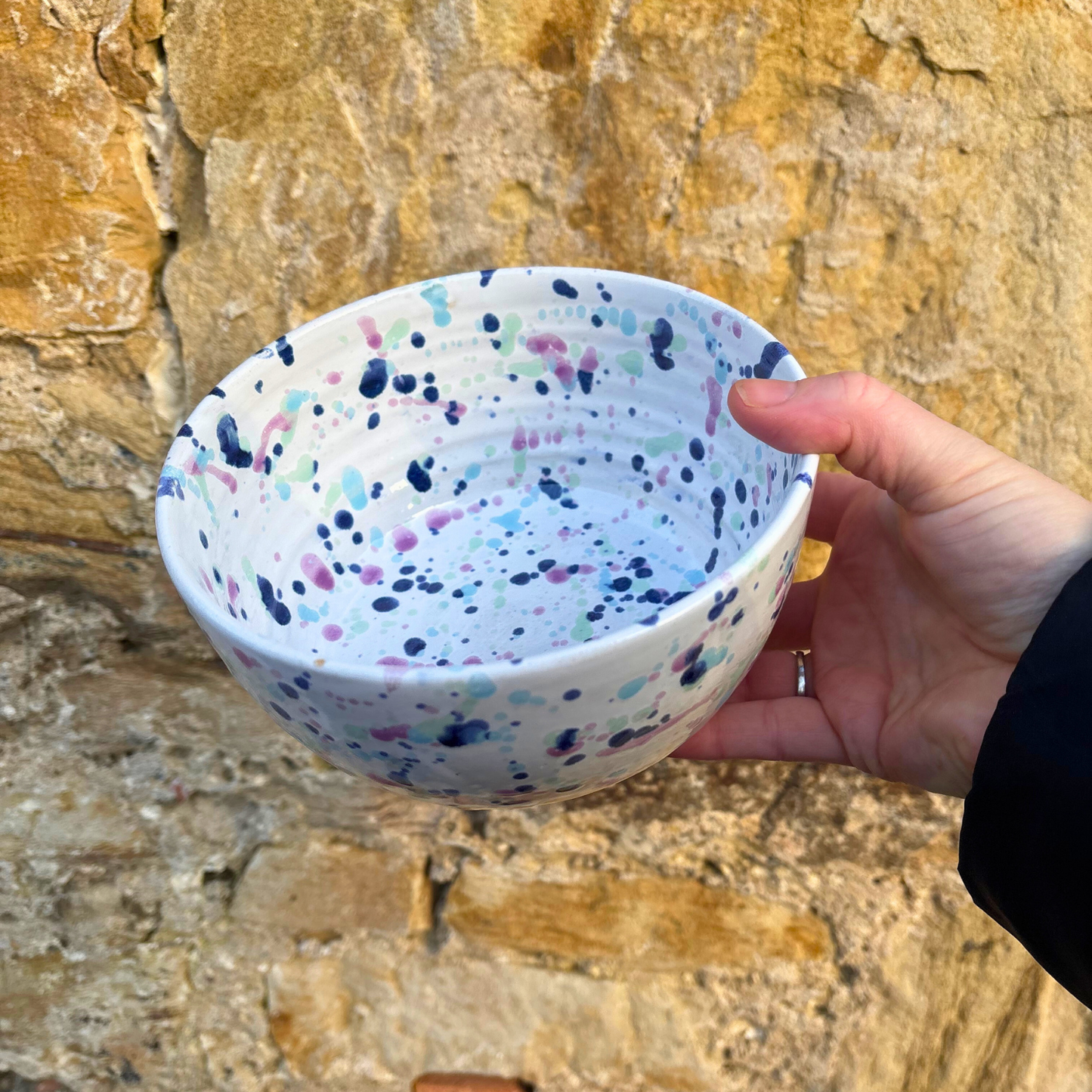 Hand holding a ceramic bowl with multicolored speckles against a stone wall background