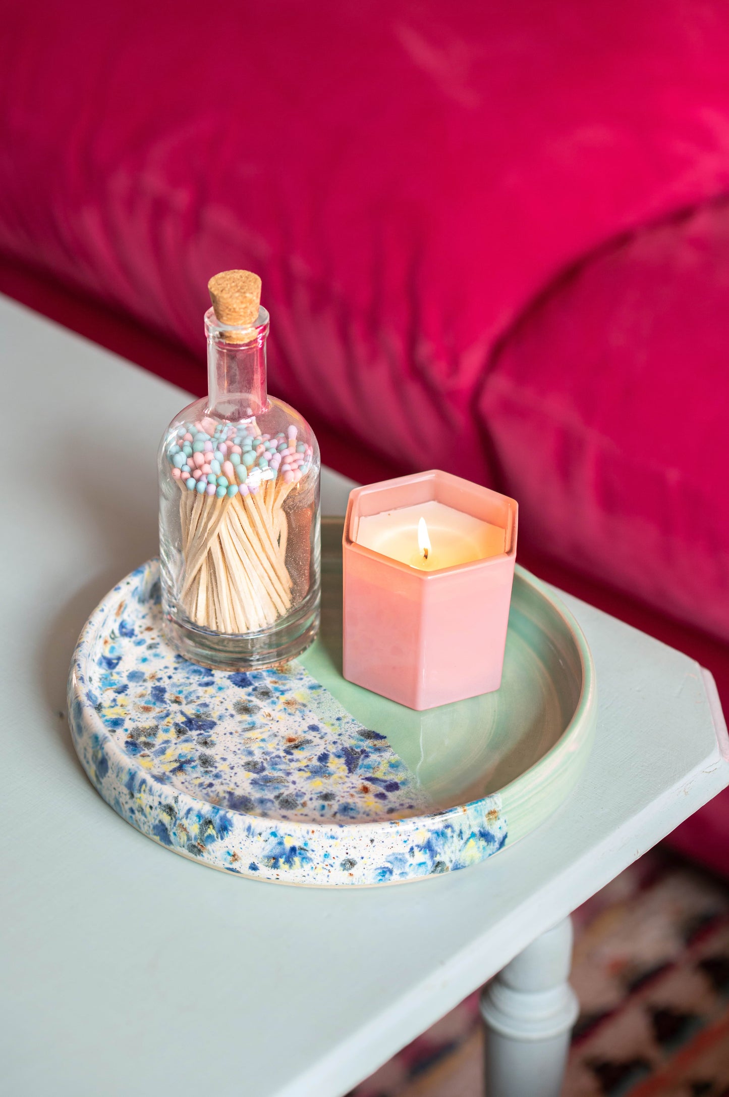 Small table with a floral-patterned tray holding a lit candle and a bottle of matches, against a pink cushioned background.