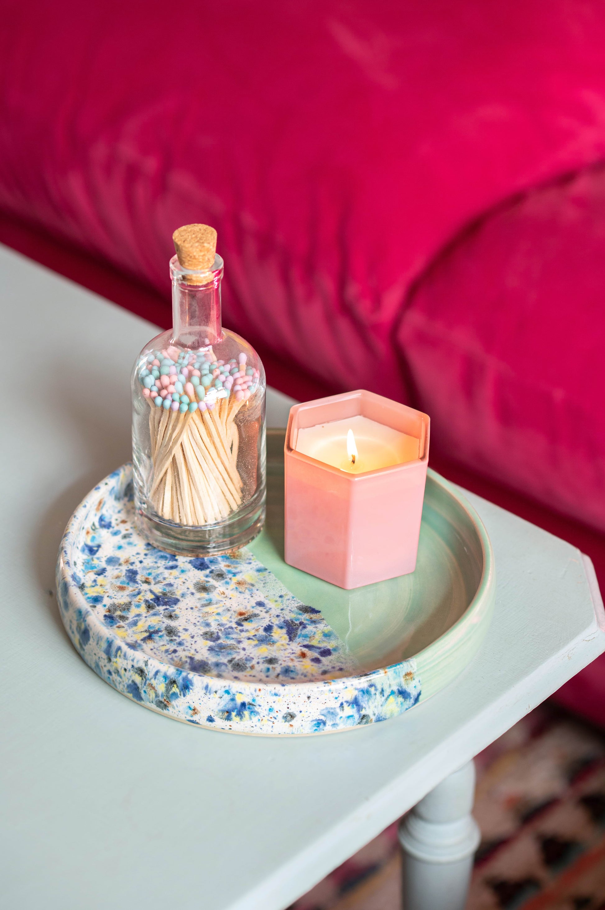 Small table with a floral-patterned tray holding a lit candle and a bottle of matches, against a pink cushioned background.