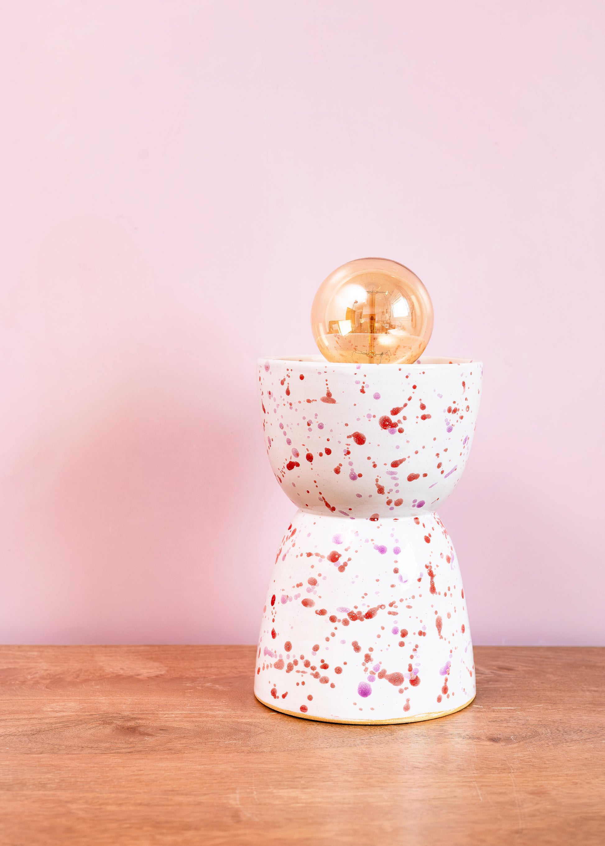 Speckled ceramic lamp with gold bulb on a wooden surface against a pink background