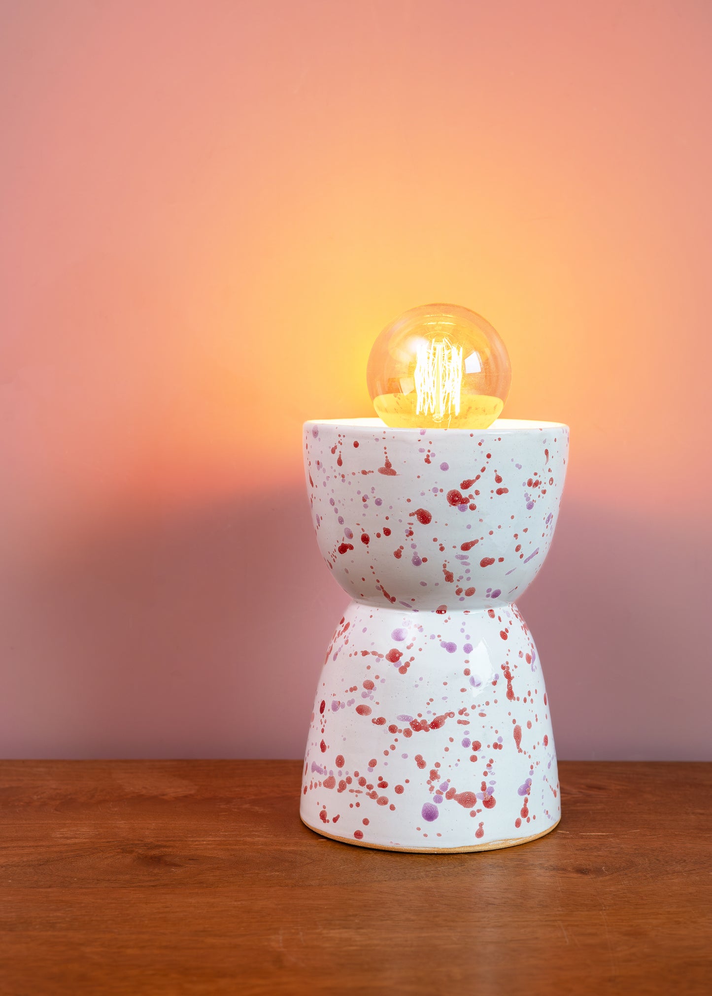 Speckled ceramic lamp with a glowing light bulb on a wooden surface and pinkish-brown background