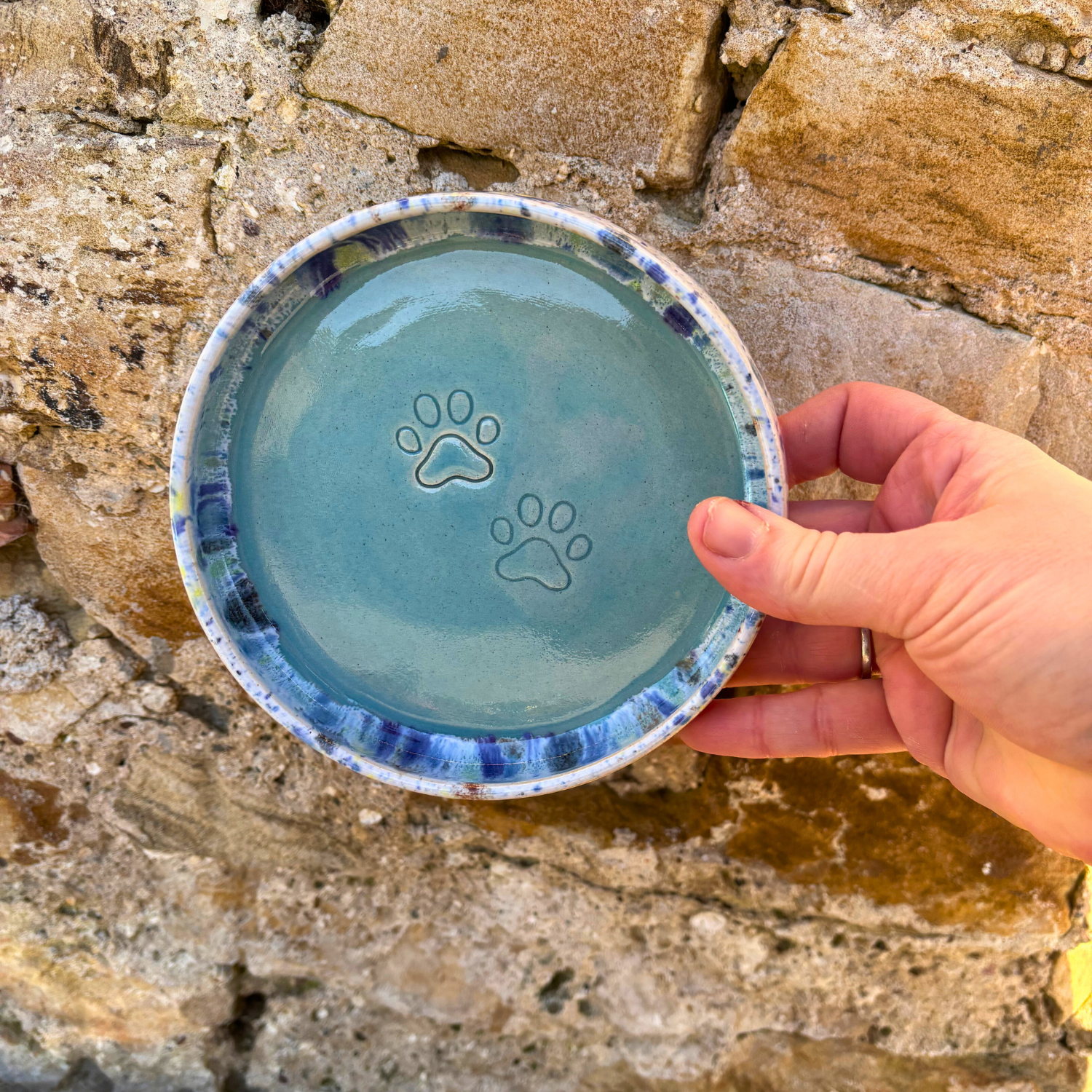 Hand holding a ceramic dish with paw prints against a stone background