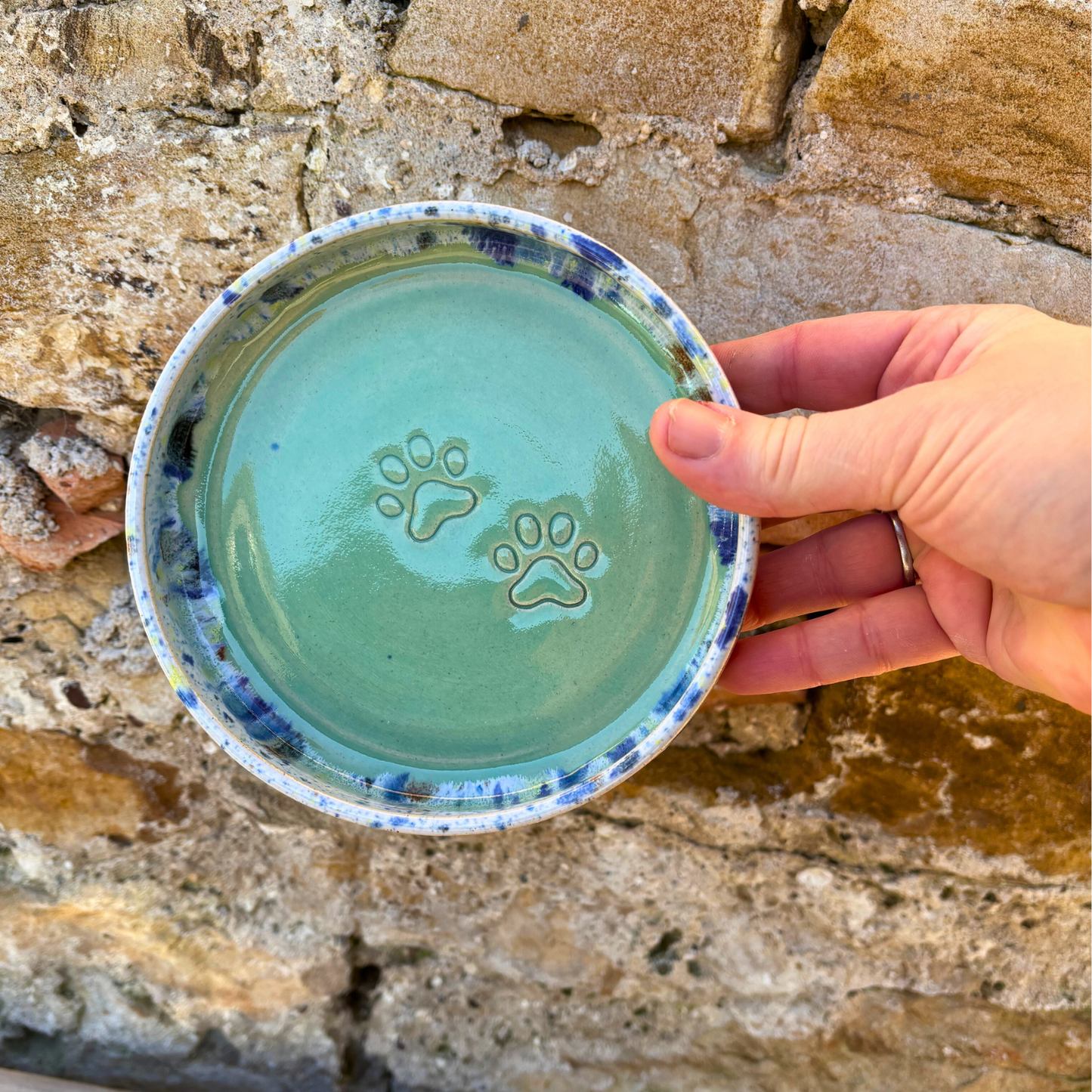 Hand holding a ceramic bowl with paw prints against a stone wall background