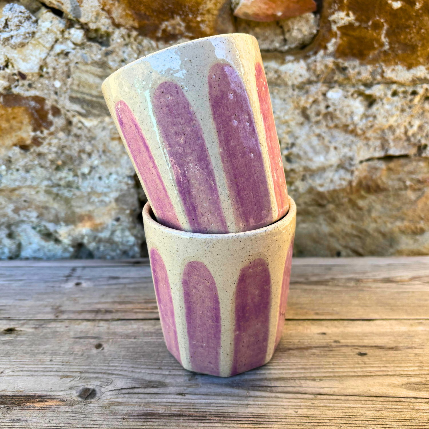 Two ceramic tumblers with striped design stacked on a wooden surface against a stone wall background