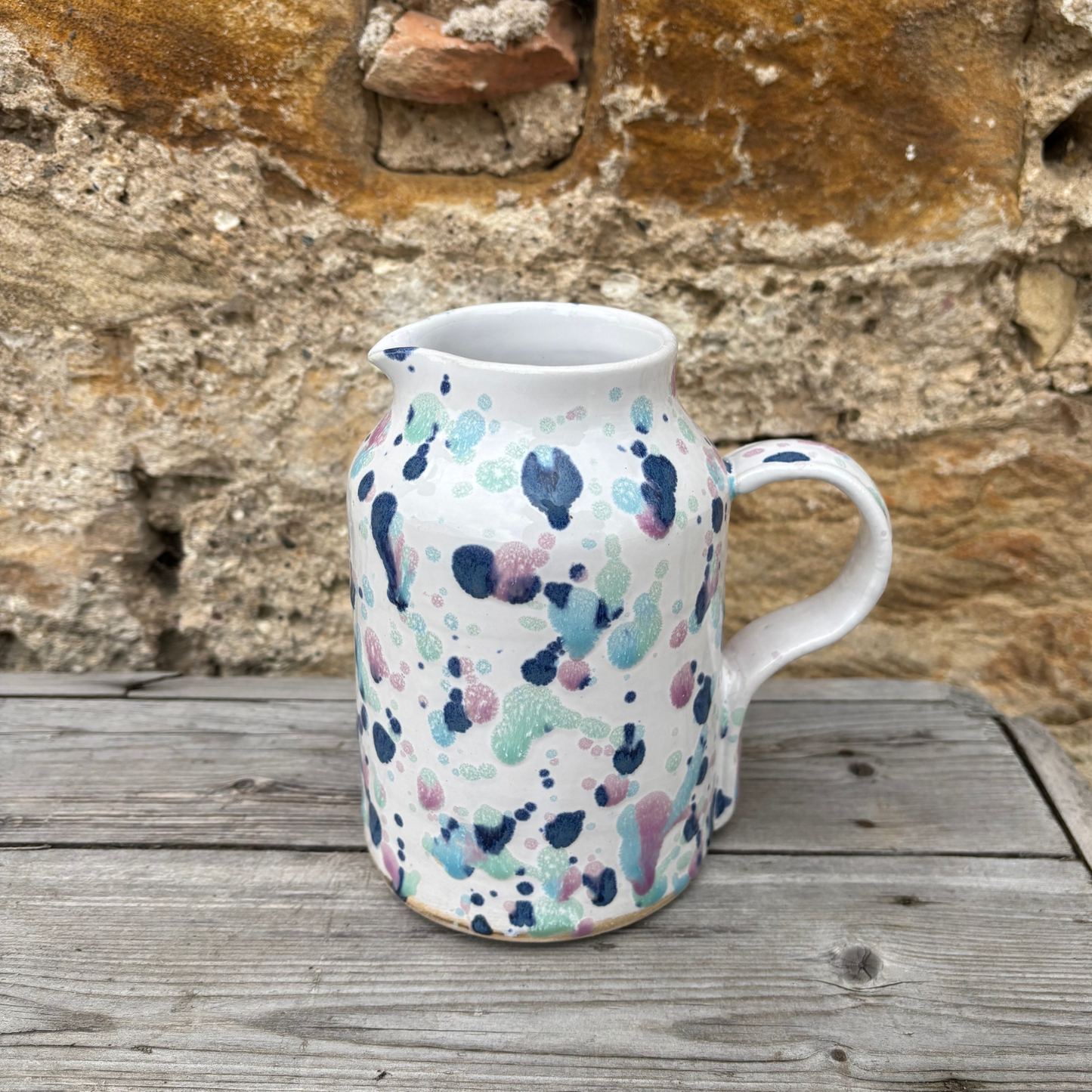 Ceramic pitcher with colorful abstract pattern on a wooden surface against a stone wall.