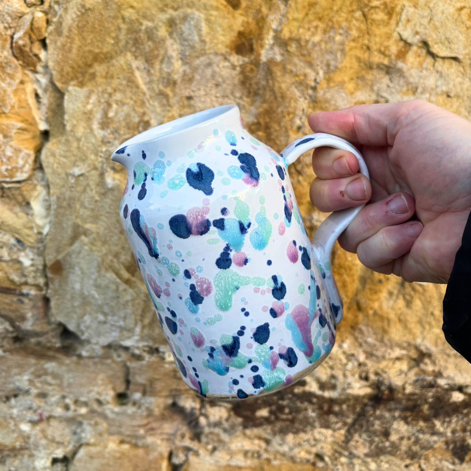 Hand holding a ceramic pitcher with colorful abstract design against a stone wall background