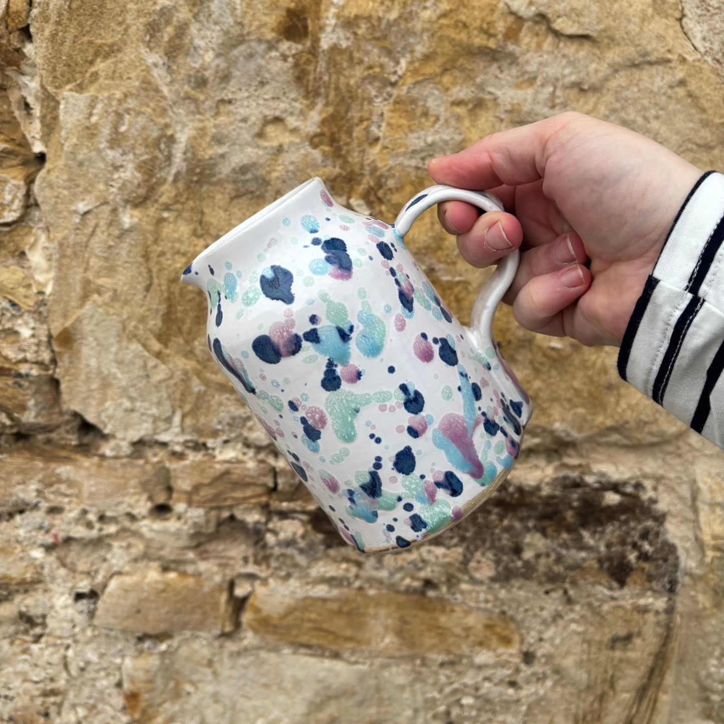 Hand holding a jug with a colorful abstract pattern against a stone wall background