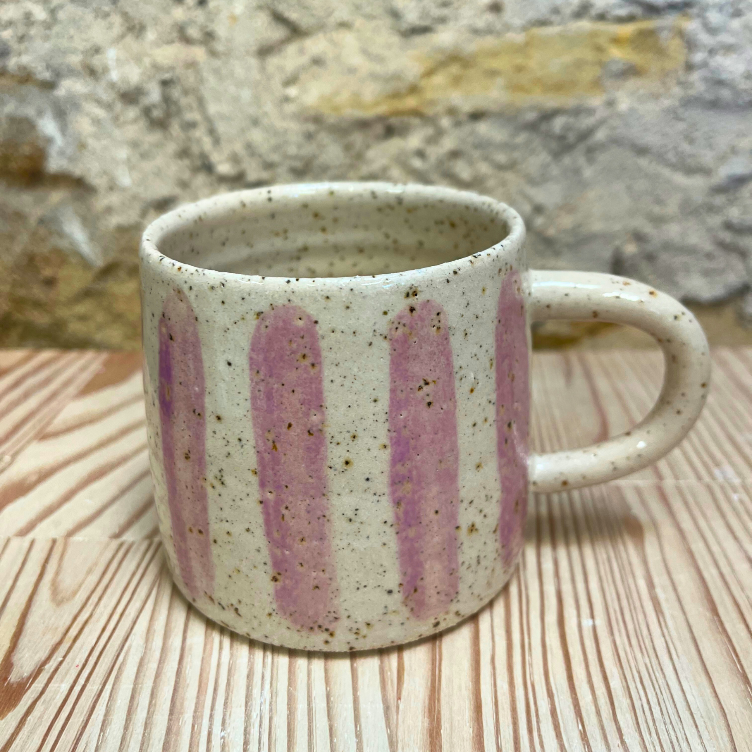 Speckled ceramic mug with pink stripes on a wooden surface with stone wall background