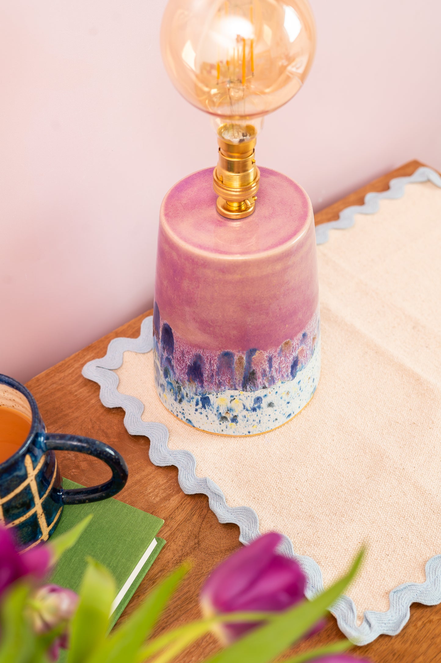 Pink ceramic lamp with a gold bulb on a wooden surface with a mug and flowers.