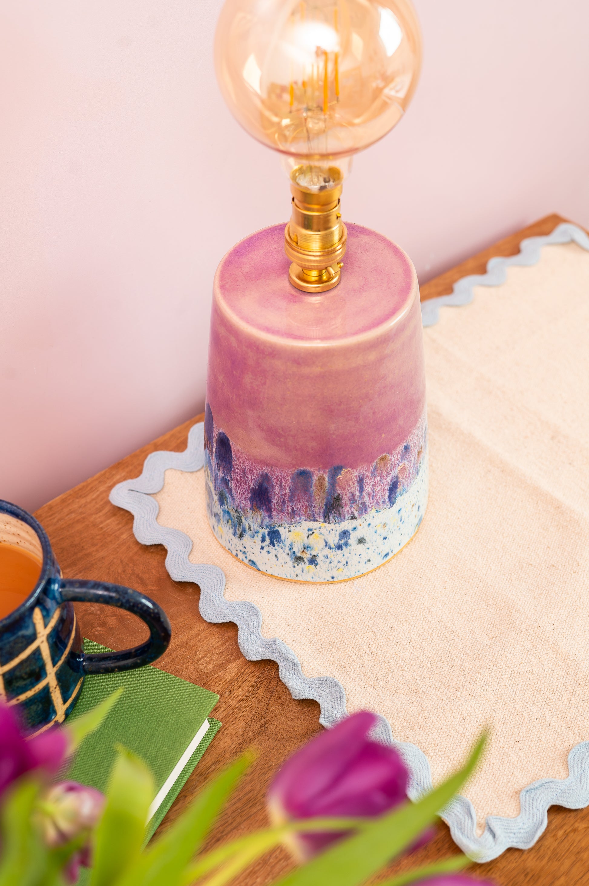 Pink ceramic lamp with a gold bulb on a wooden surface with a mug and flowers.
