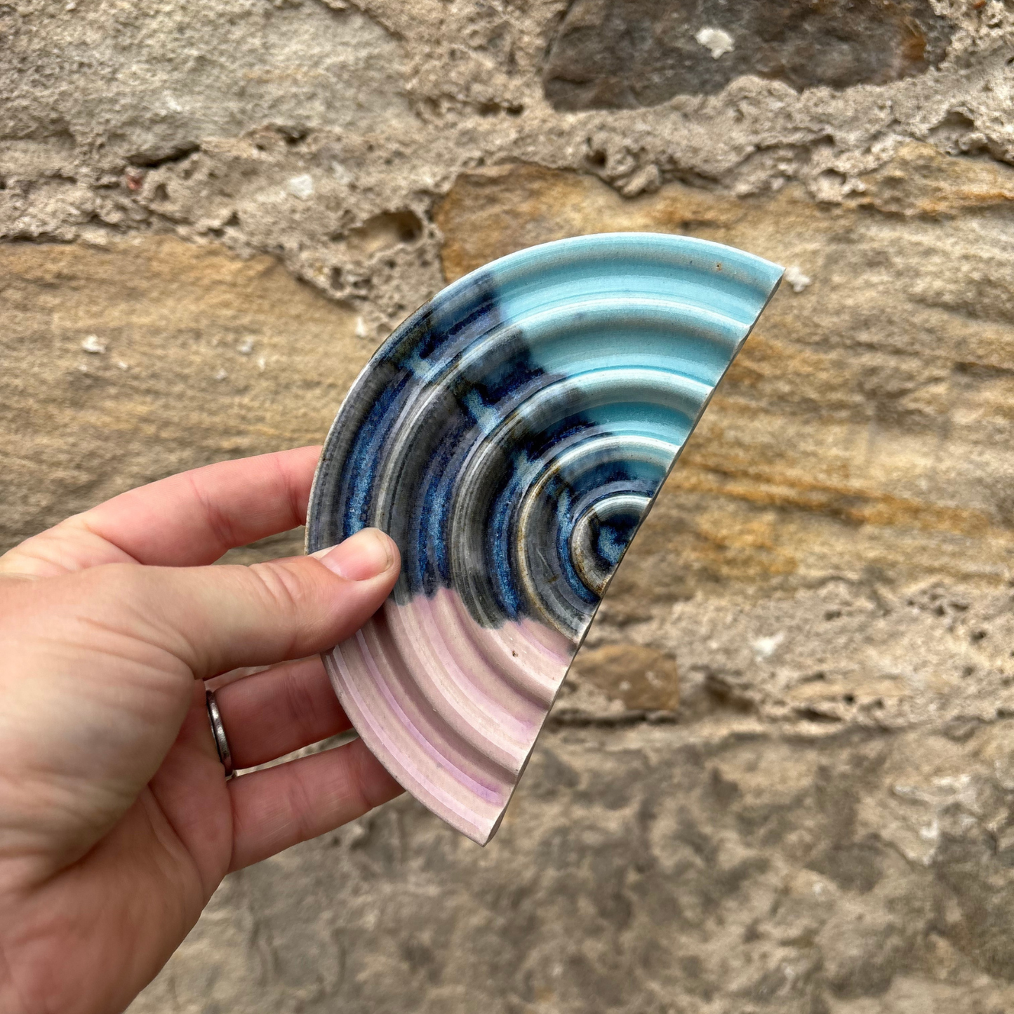 Hand holding a ceramic soap dish with blue and pink patterns against a stone wall background