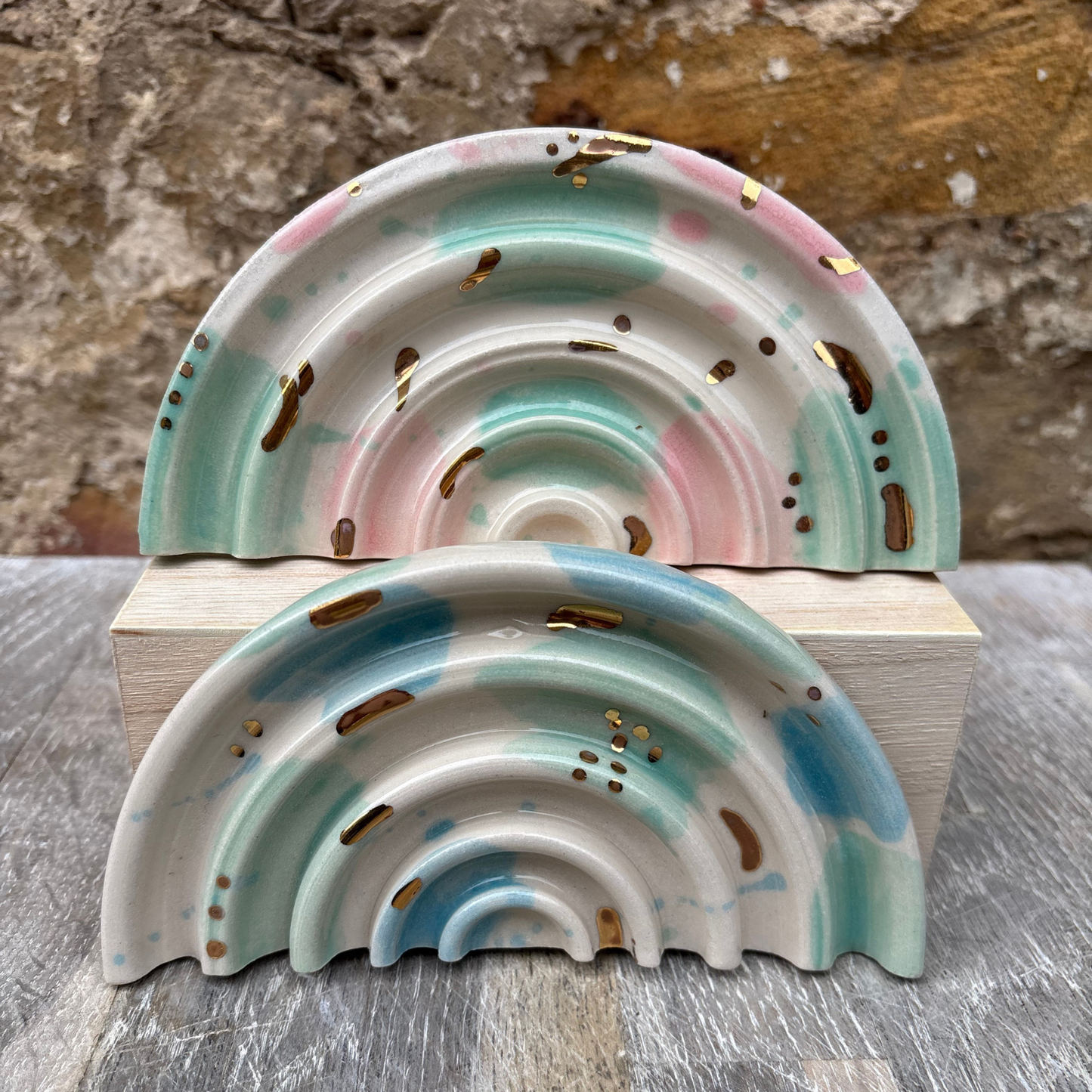 Two ceramic rainbow-shaped soap dishes with gold speckles on a wooden block against a stone wall background.