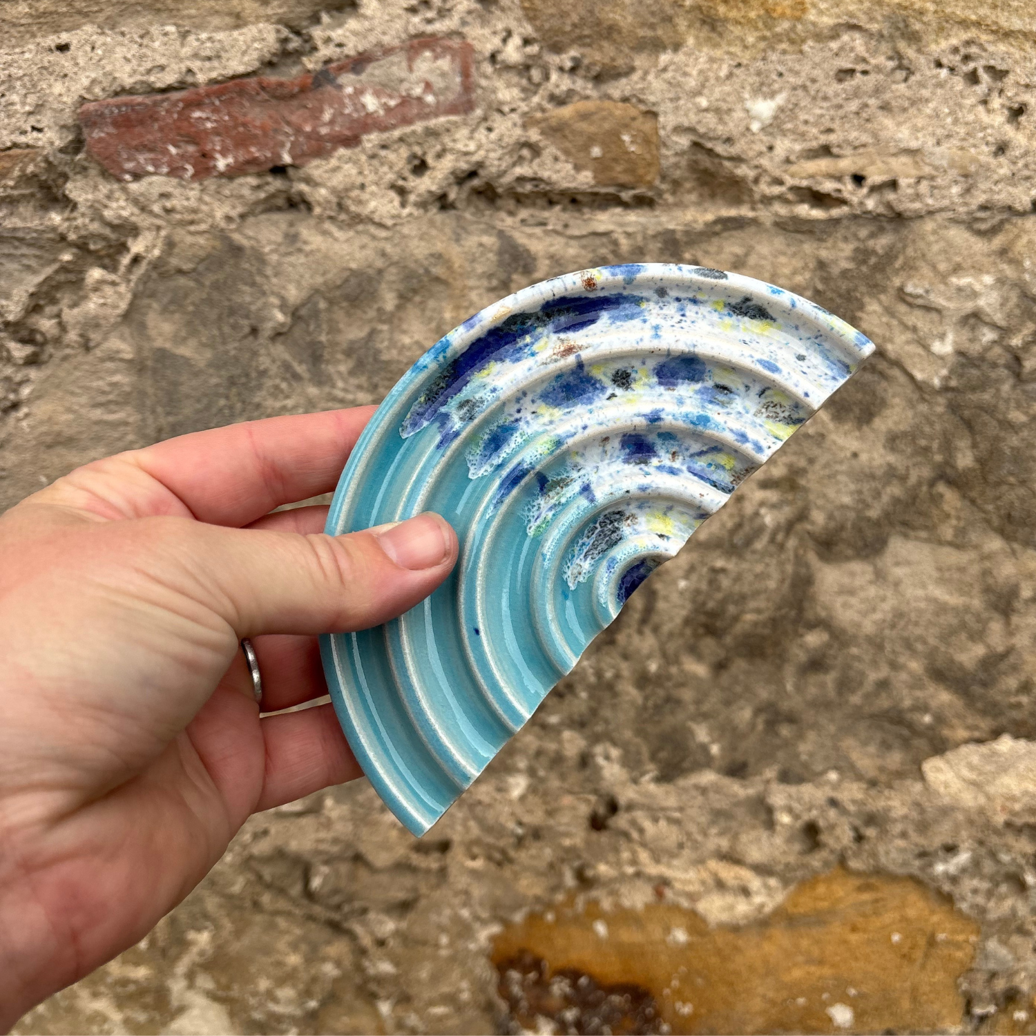 Hand holding a ceramic soap dish with blue and white patterns against a stone wall background