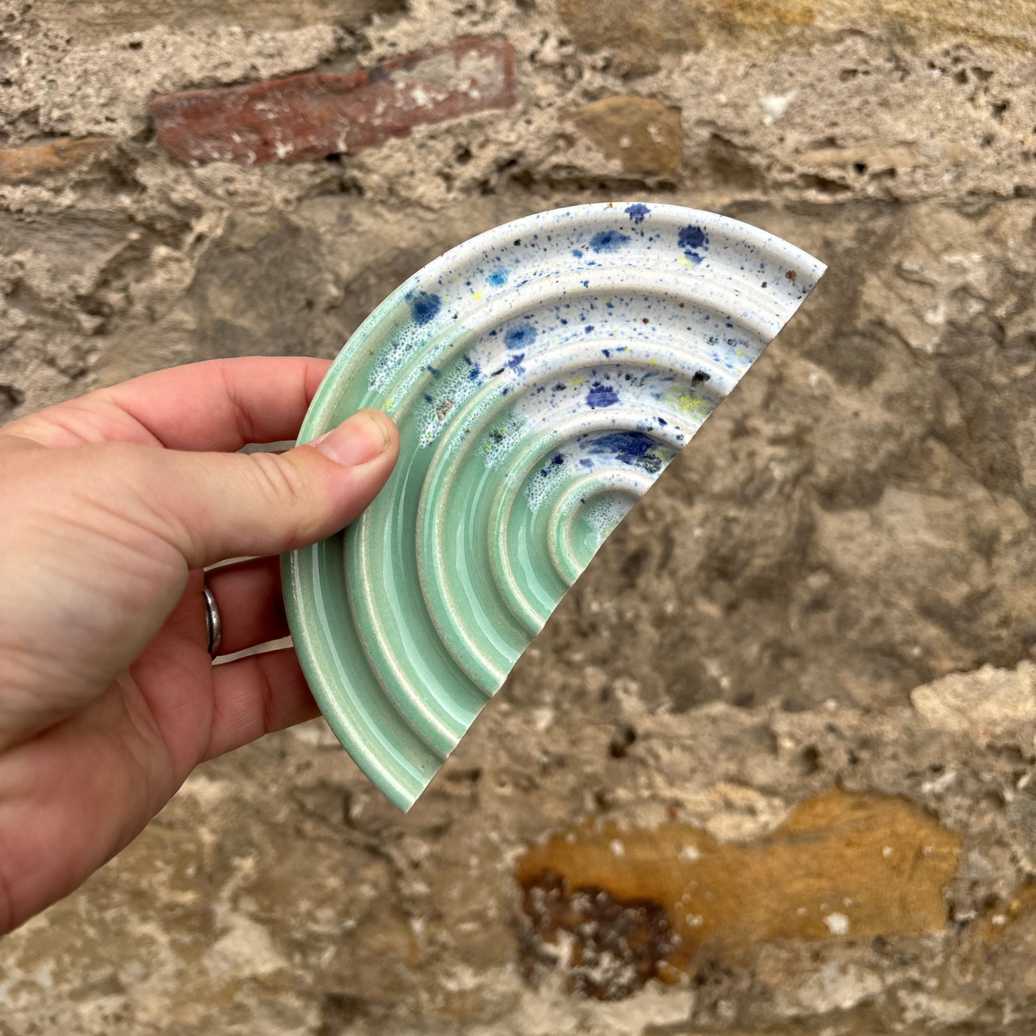 Hand holding a ceramic soap dish with wave design against a stone wall background