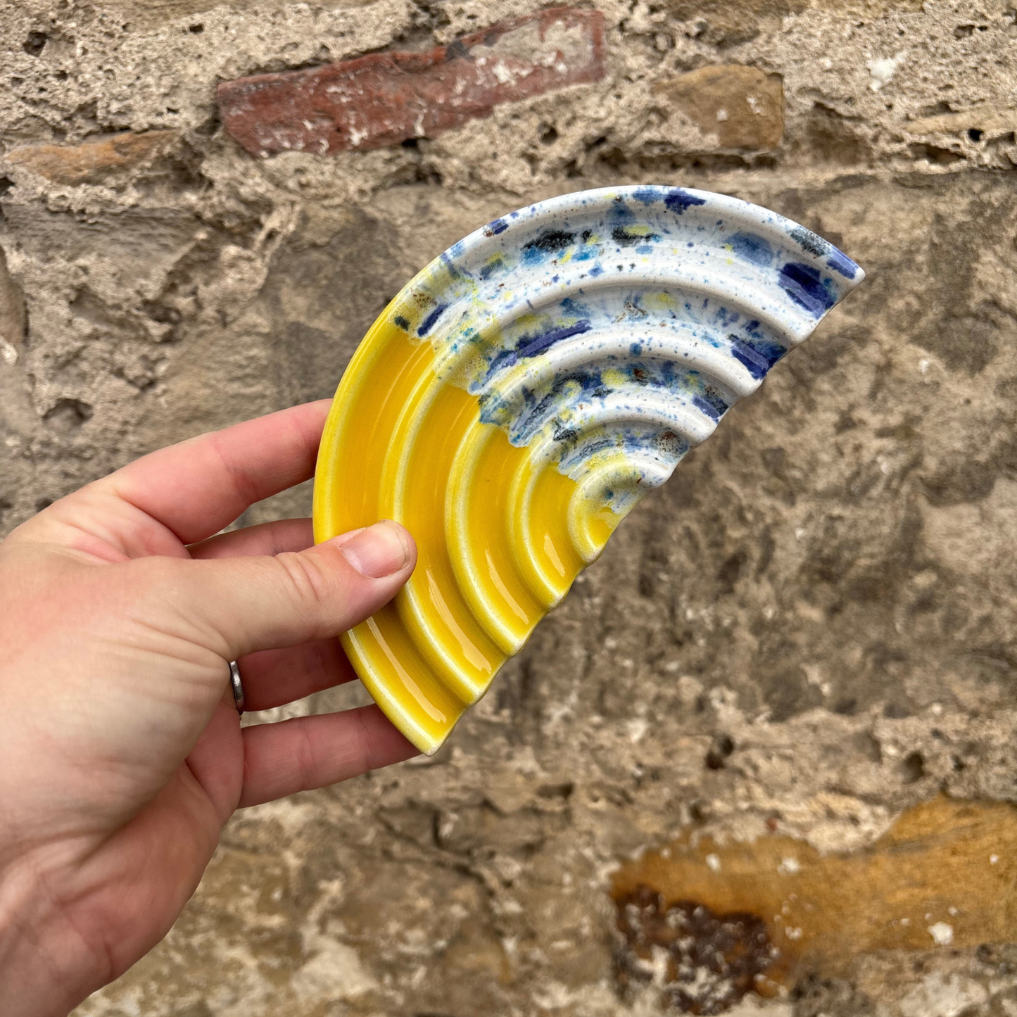Hand holding a ceramic soap dish with yellow and blue design against a stone wall background