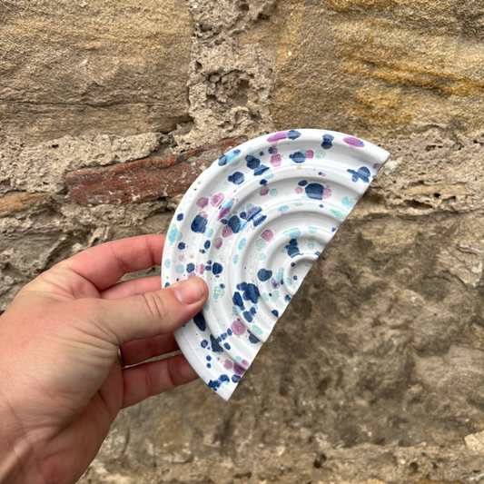 Hand holding a ceramic soap dish with colorful patterns against a stone wall background
