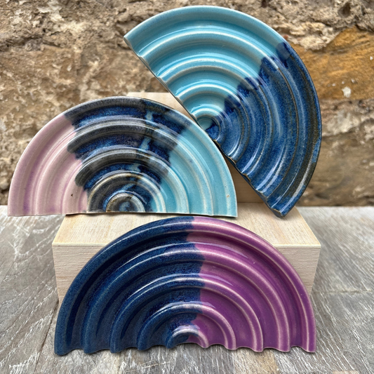 Three ceramic plates with blue and purple glaze on a wooden block against a stone wall background.