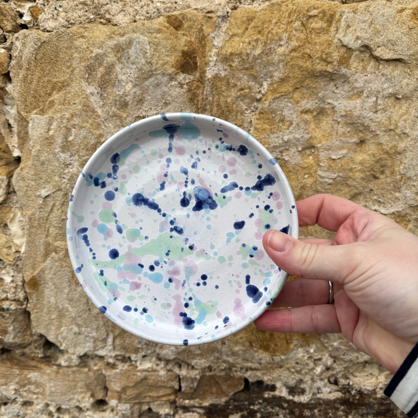 Hand holding a small ceramic plate with colorful splatter design against a stone wall background