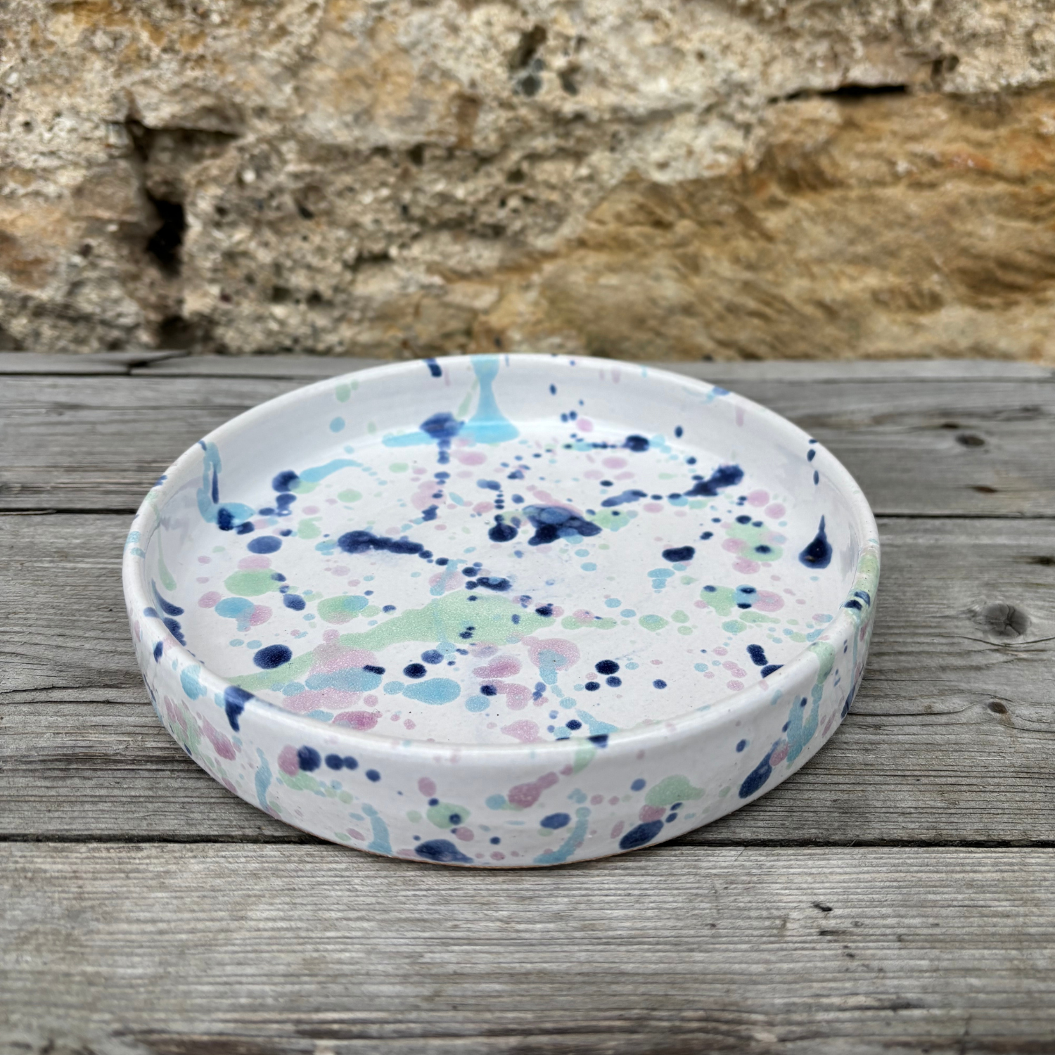 Round ceramic dish with colorful splatter pattern on a wooden surface with stone background
