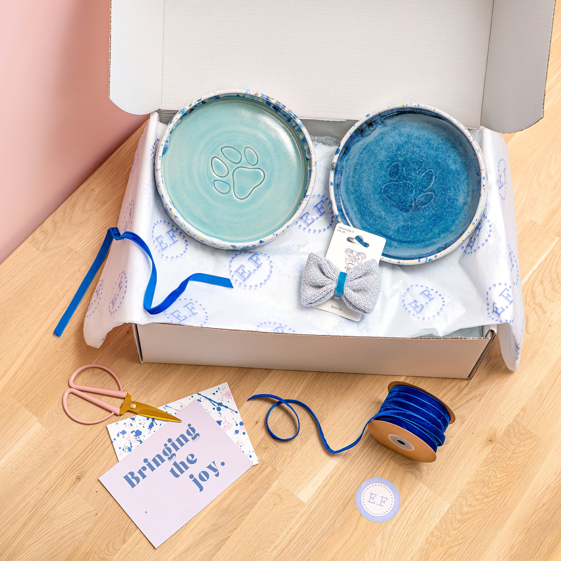 Two ceramic pet bowls with paw prints, a bow tie, and craft supplies in a box on a wooden surface.