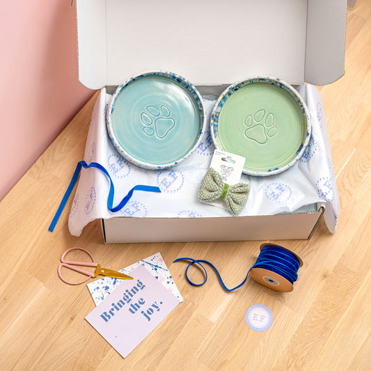 Two ceramic dog bowls with paw prints, a bow tie, and packaging on a wooden surface.