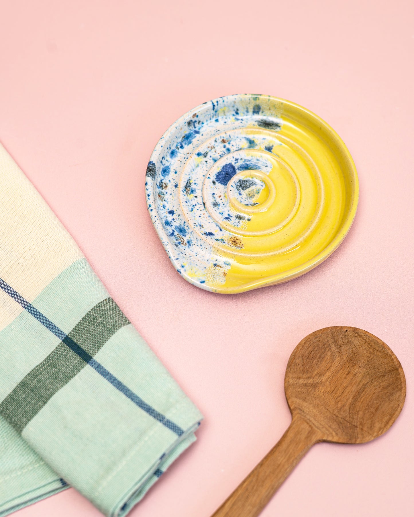 Ceramic plate with blue and yellow swirl design, wooden spoon, and green checkered towel on a pink background
