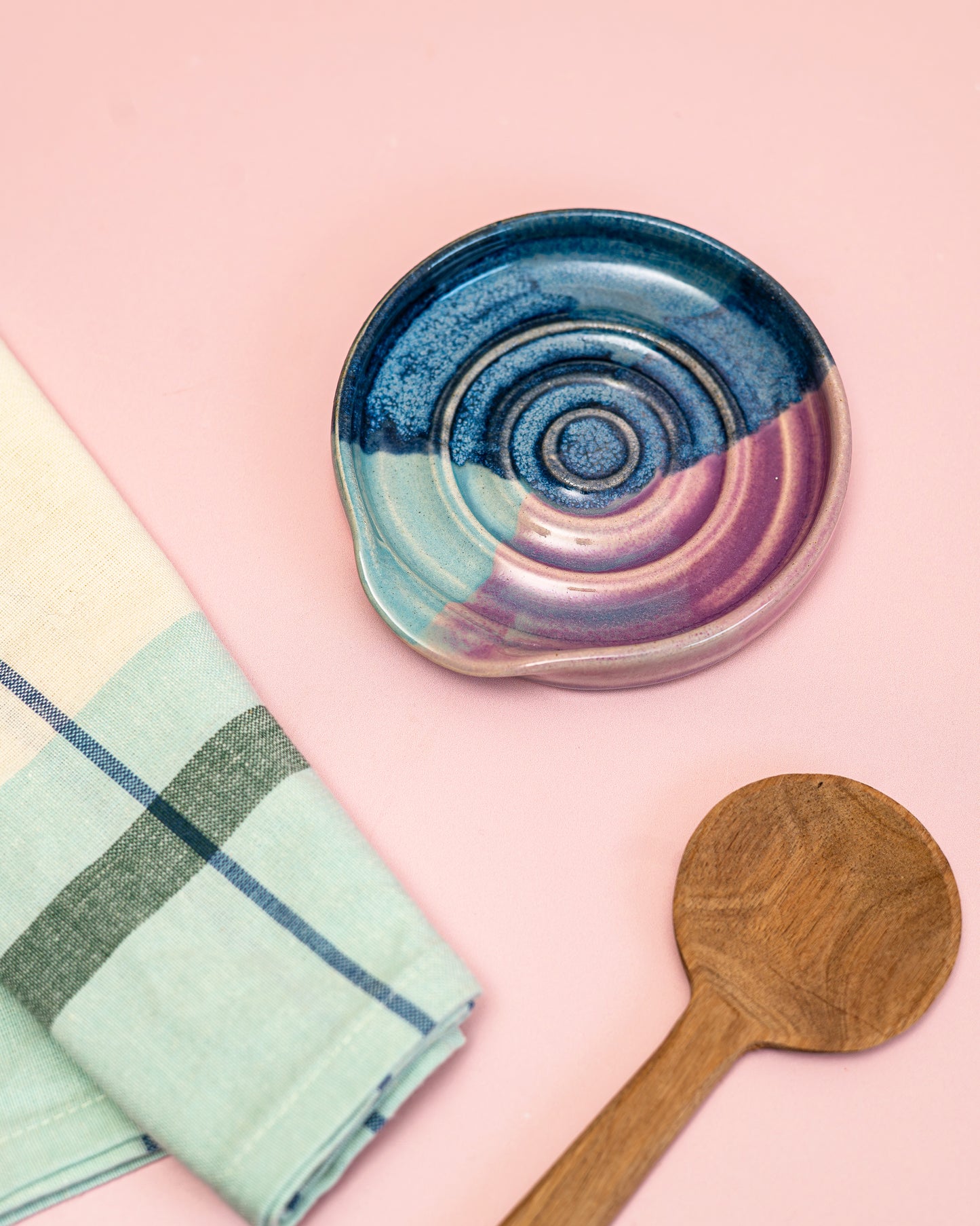 Ceramic dish with concentric circle pattern, wooden spoon, and plaid towel on a pink background