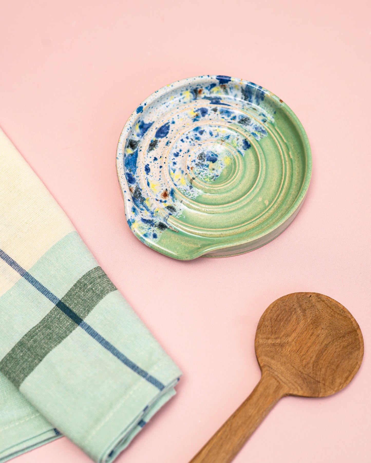 Green ceramic dish with blue and yellow speckles, wooden spoon, and plaid towel on a pink background