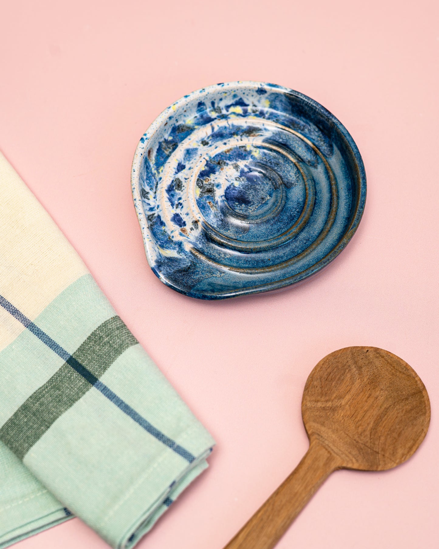 Blue ceramic dish, green checkered towel, and wooden spoon on a pink background