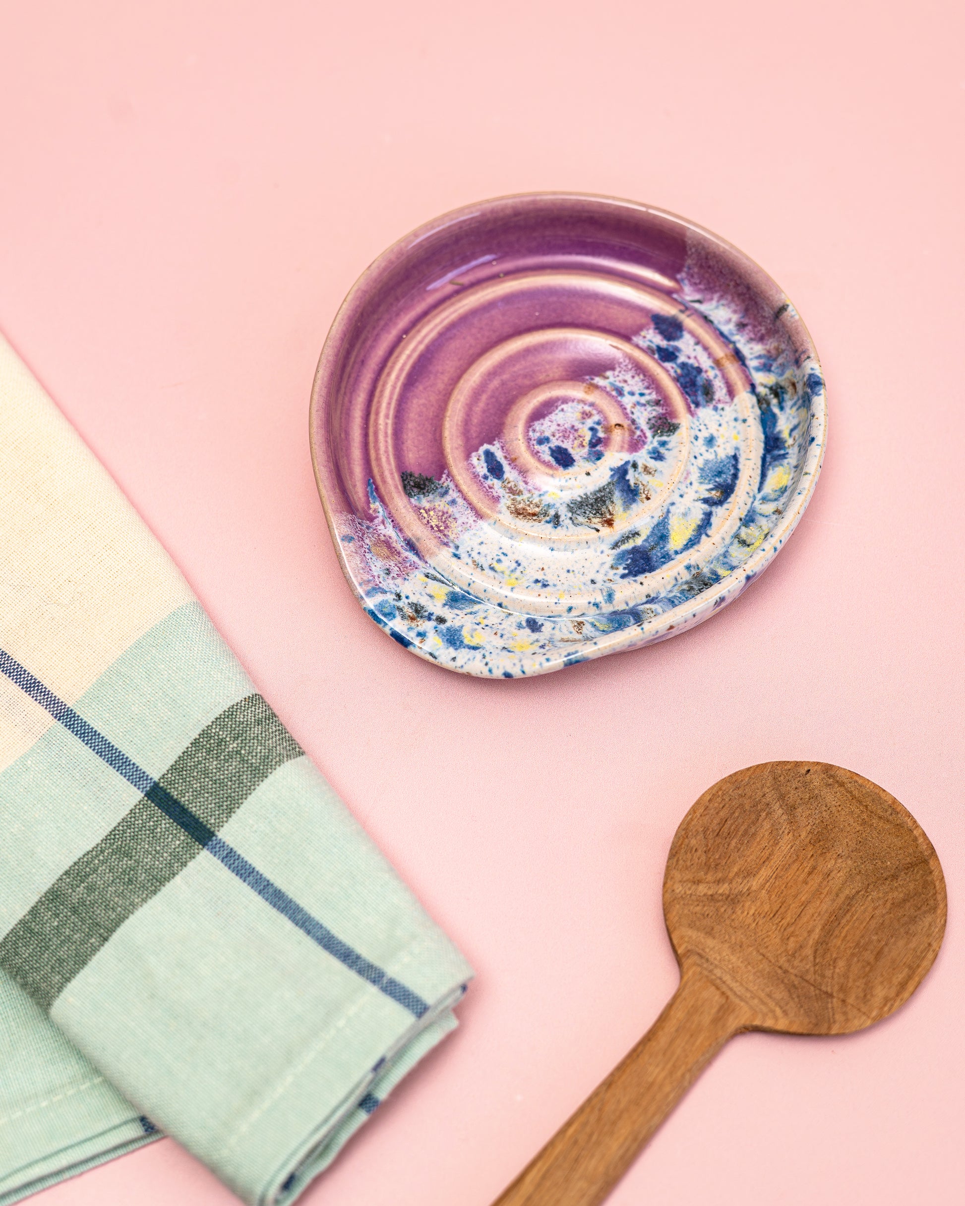 Ceramic plate with a colorful spiral design on a pink background, accompanied by a wooden spoon and green checkered towel.