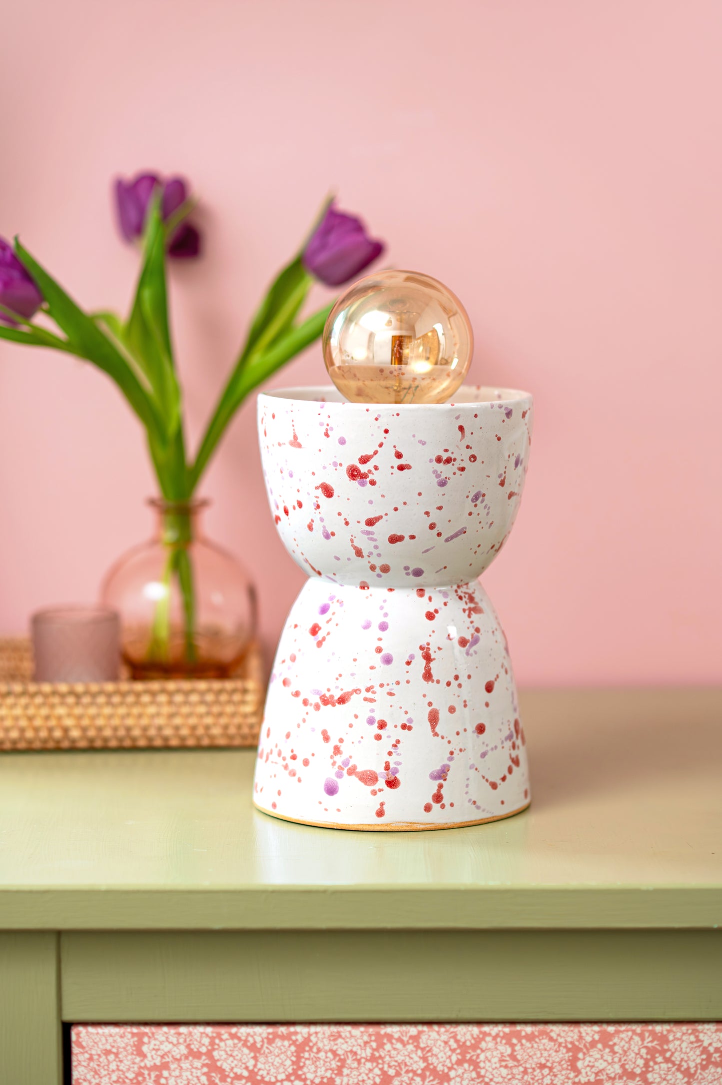 Speckled ceramic lamp on a table with a pink wall and flowers in the background