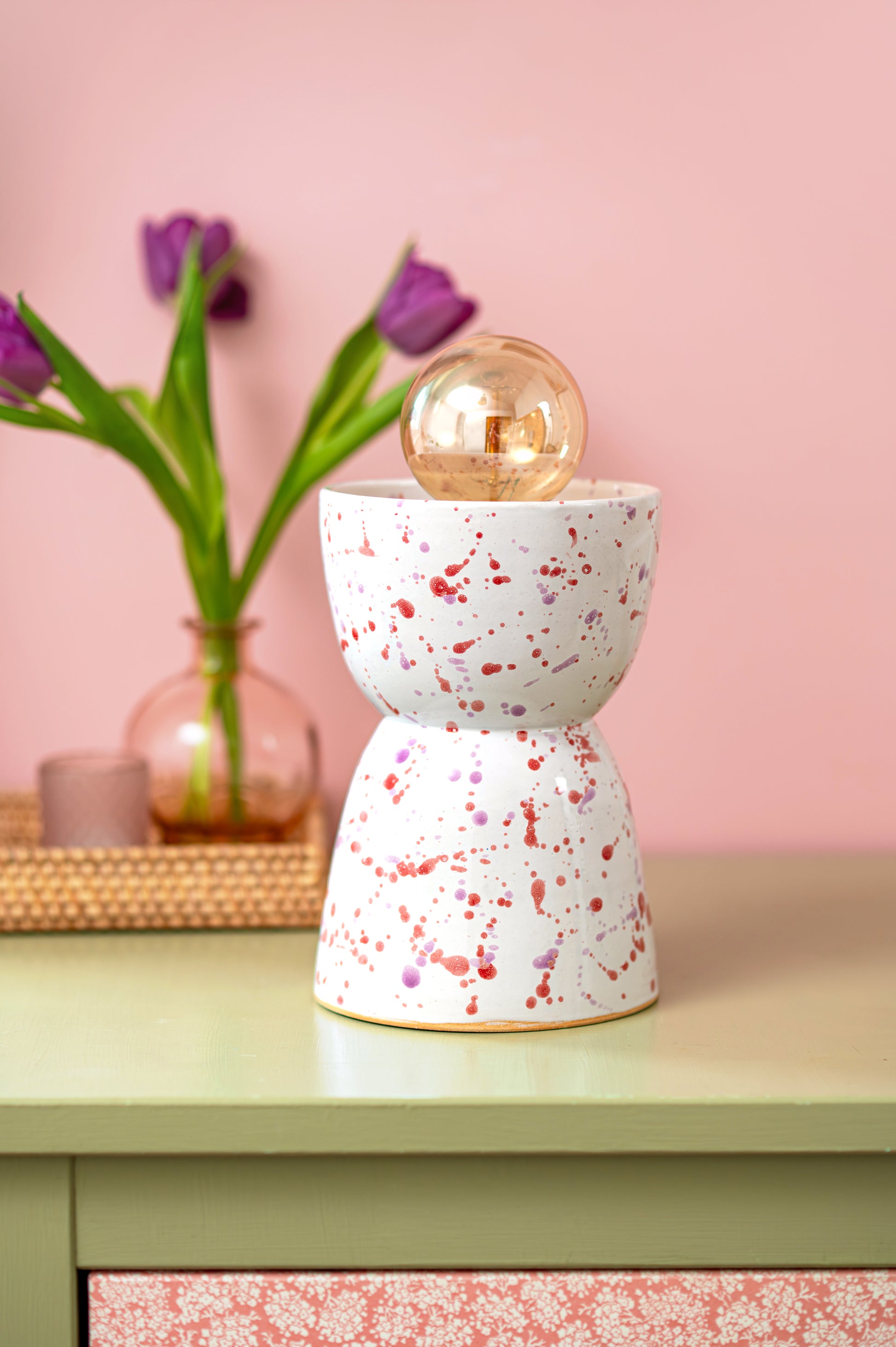 Speckled ceramic lamp on a table with a pink wall and flowers in the background