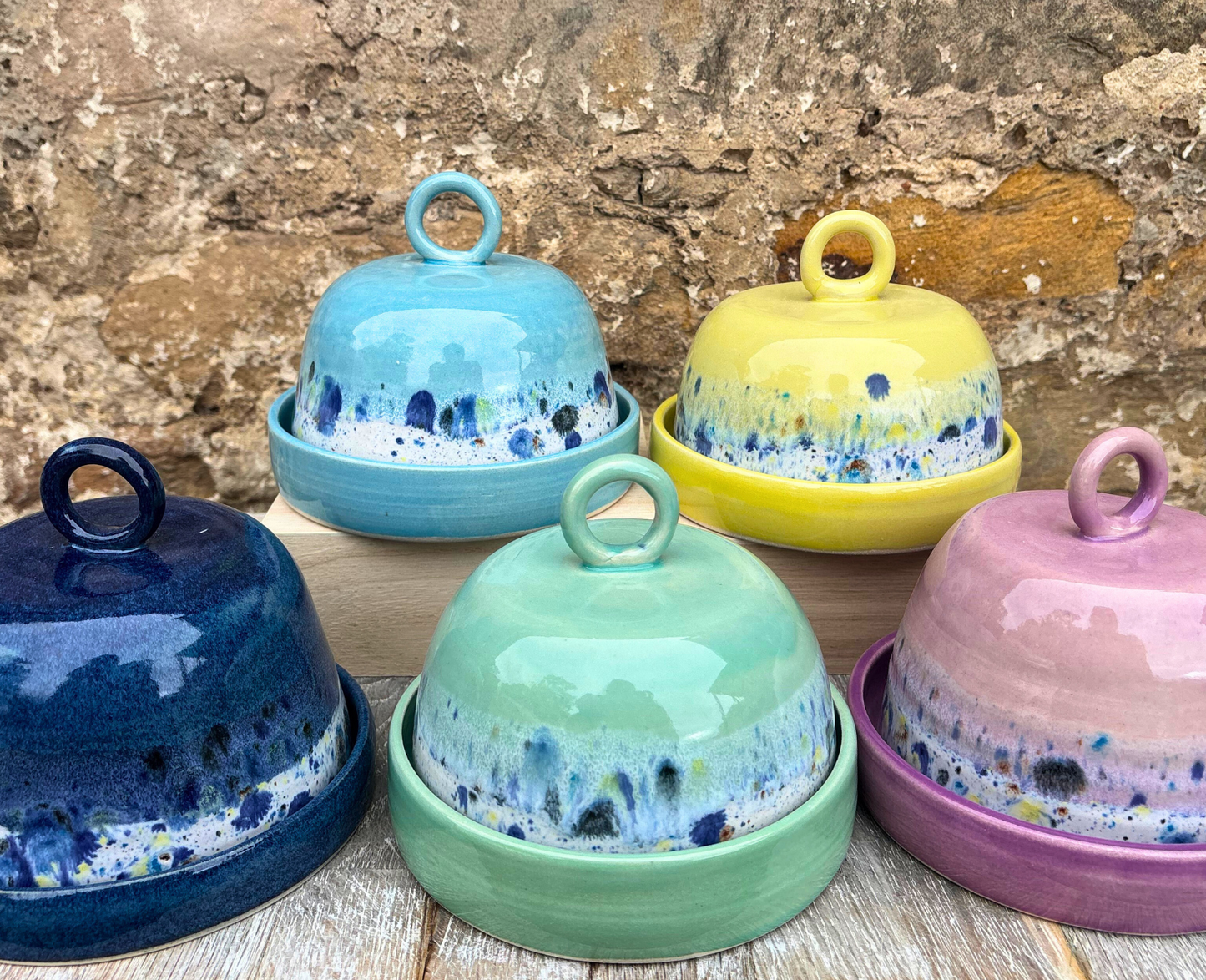 Set of colorful ceramic covered dishes on a wooden surface with a stone wall background.