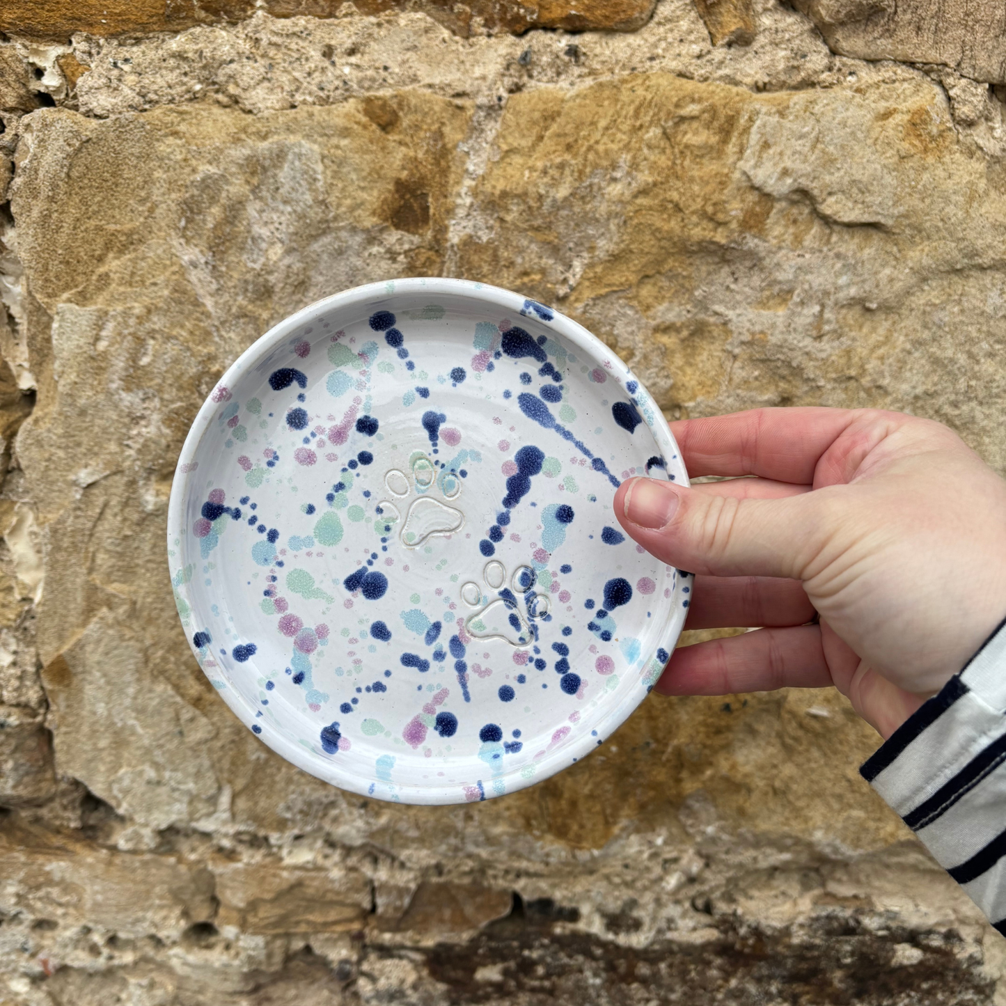Hand holding a small ceramic plate with blue and purple splatter design against a stone wall background