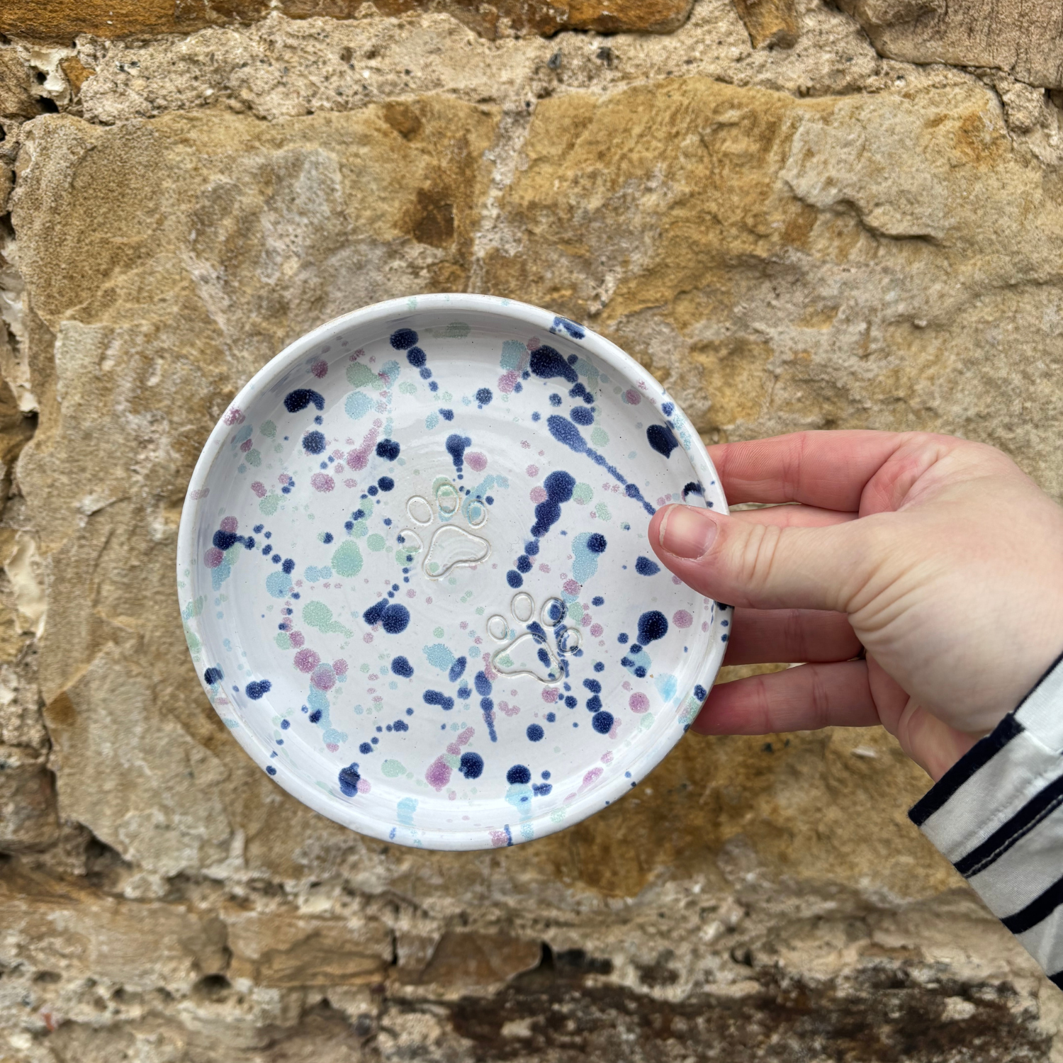 Hand holding a small ceramic plate with blue and purple splatter design against a stone wall background