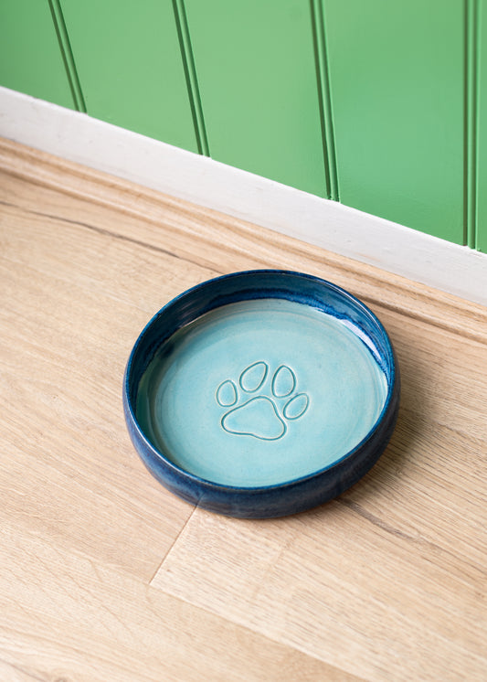Blue ceramic pet bowl with paw print design on a wooden surface against a green wall.